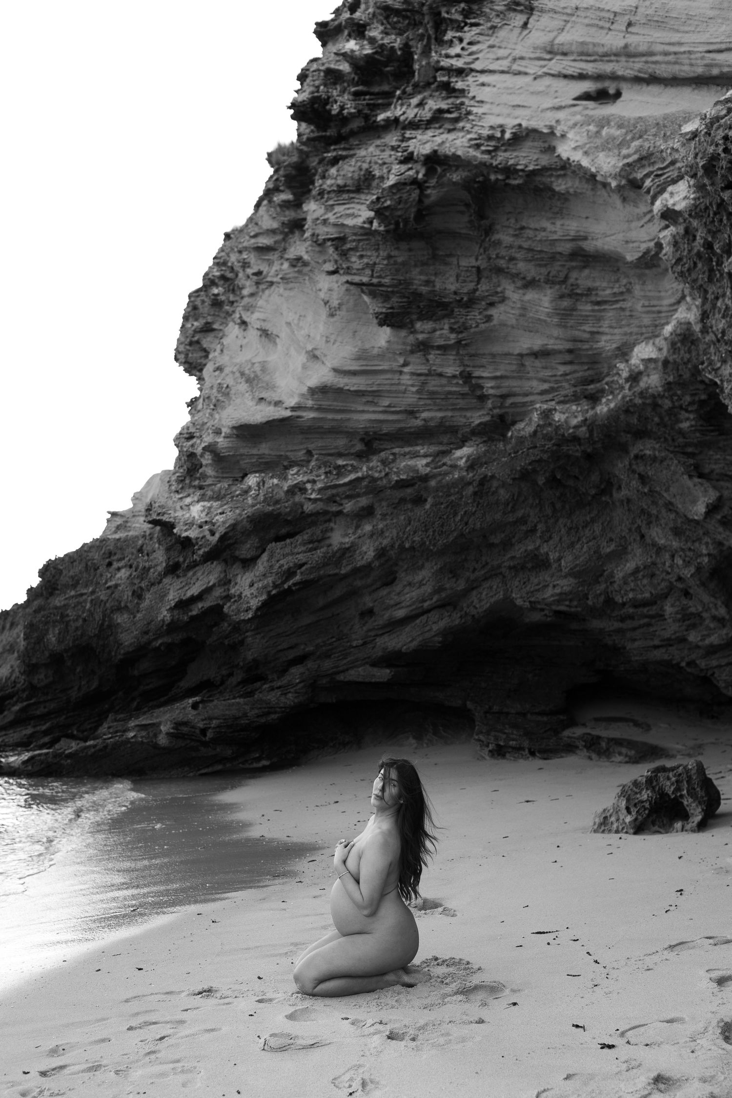 A black and white photo of someone sitting on a sandy beach beneath dramatic rocky cliffs.