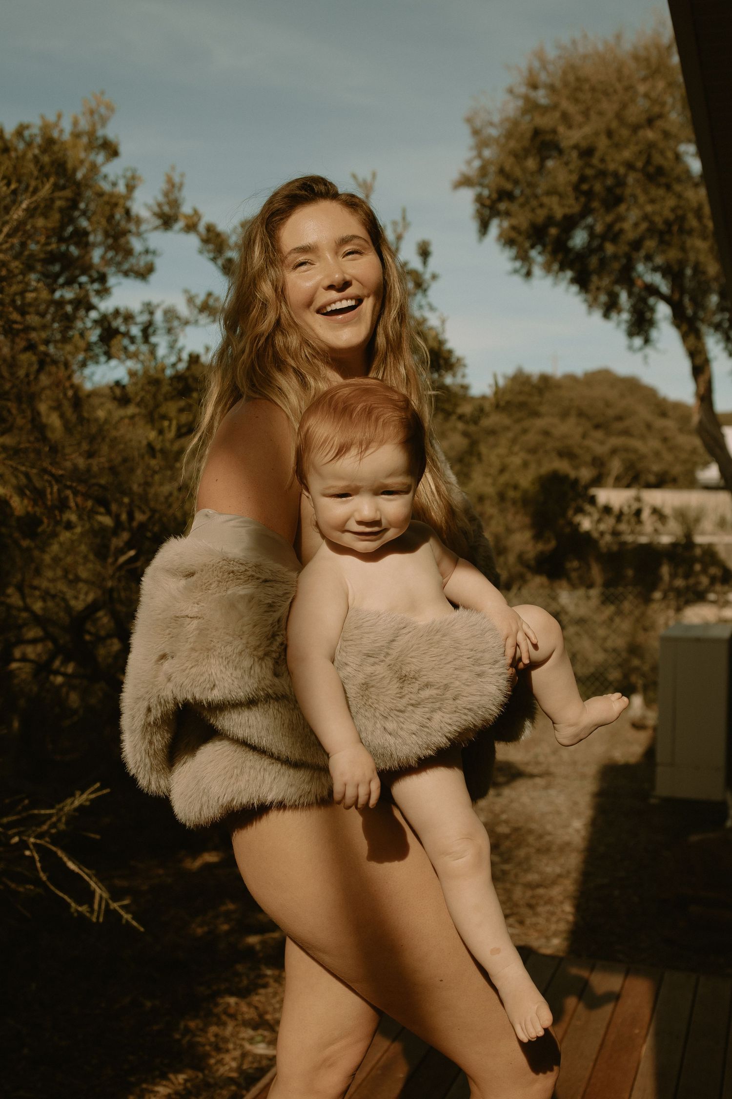Candid outdoor portrait in warm sunlight showing joyful moment between adult and baby wrapped in cozy beige blanket.