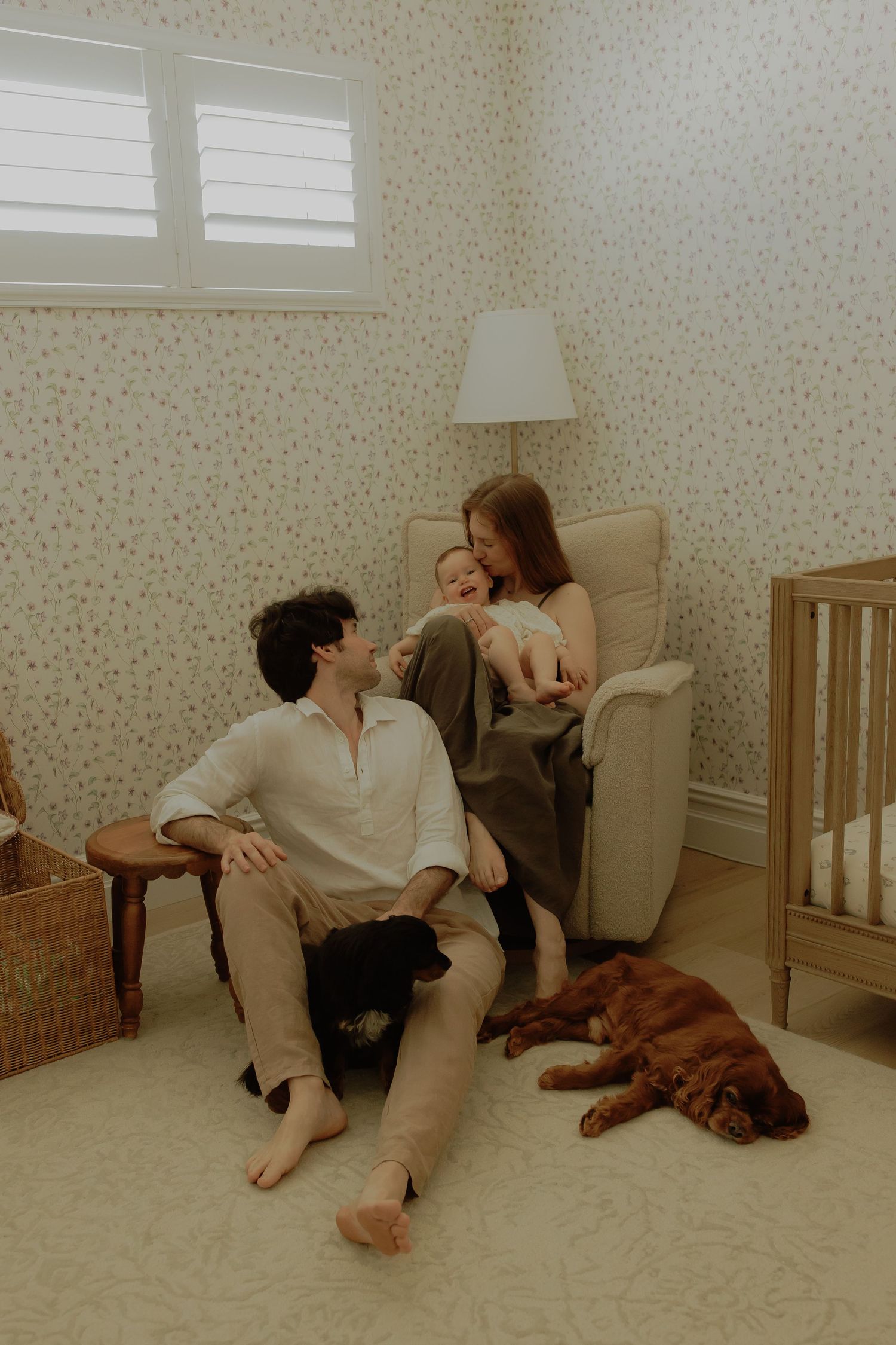 A young family with a baby and two dogs relaxes in a cozy nursery with floral wallpaper and neutral decor.