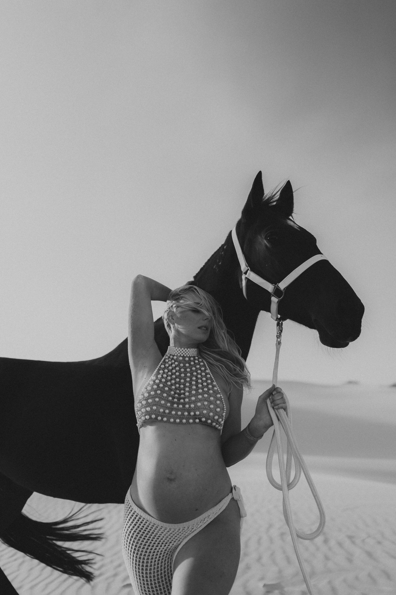 Black and white photo of a blonde woman in a bikini holding a horse's lead rope in a sandy desert landscape.