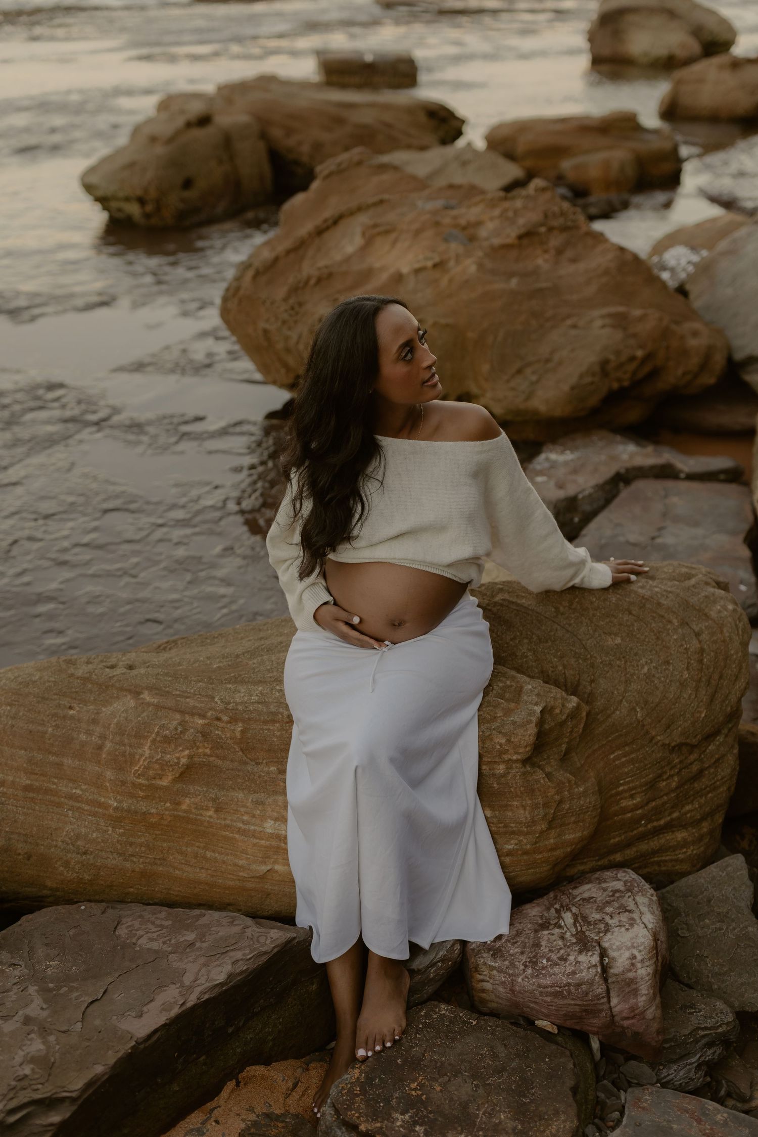 Pregnant woman in white crop top and skirt poses on coastal rocks at dusk.