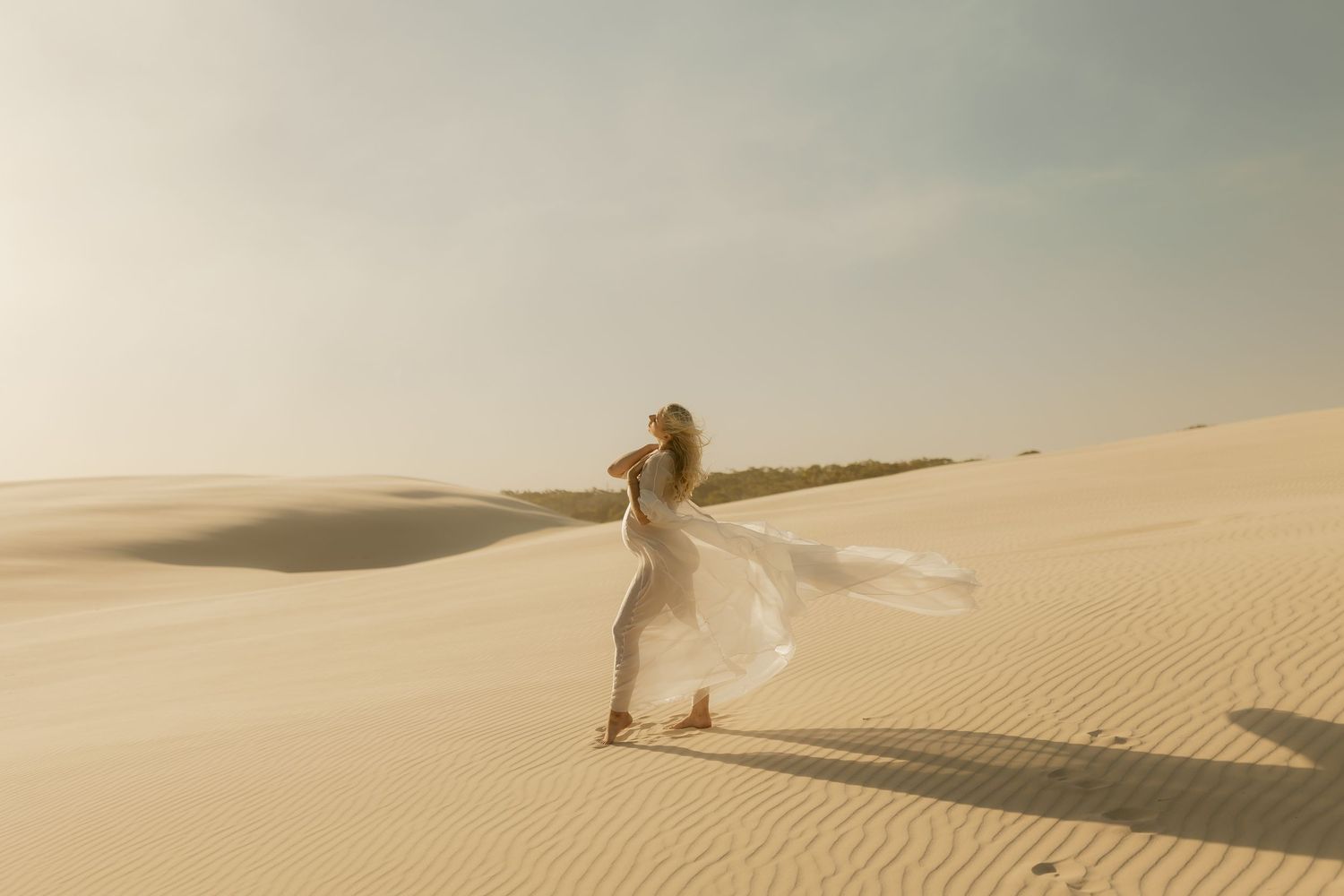 Woman in flowing white dress standing on sand dunes, dress blowing in wind against a soft desert sky.