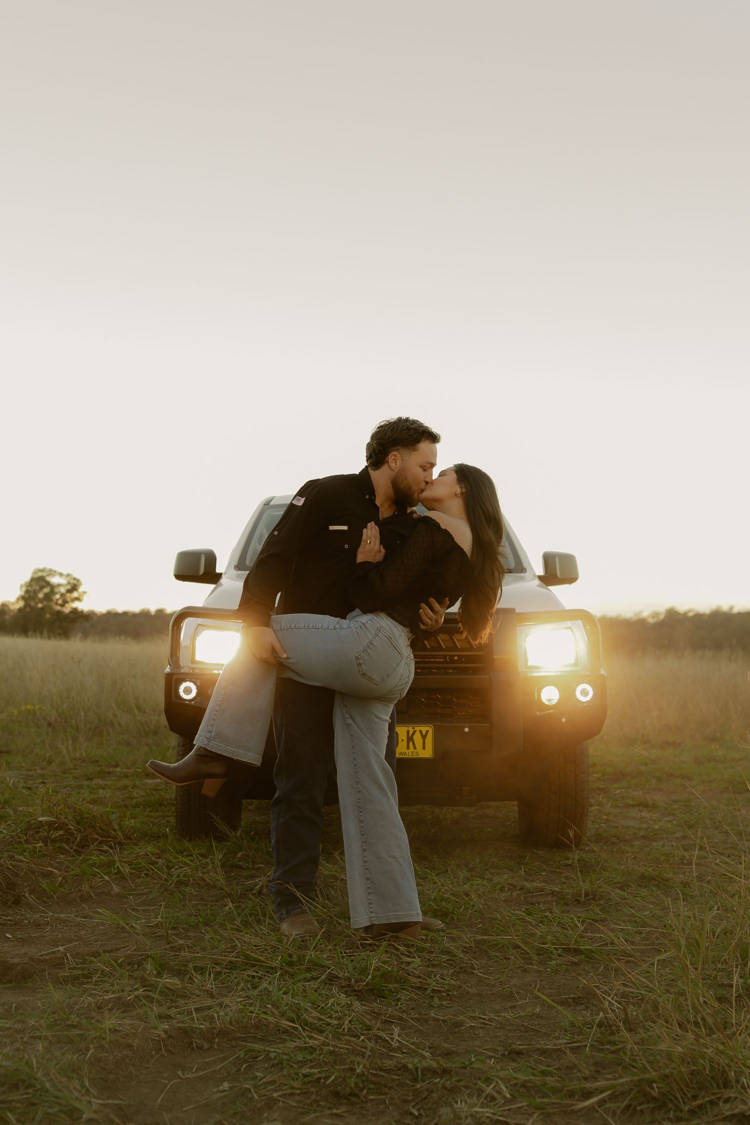 Couple sharing a romantic kiss in front of a 4WD vehicle with headlights on in an open field at sunset.