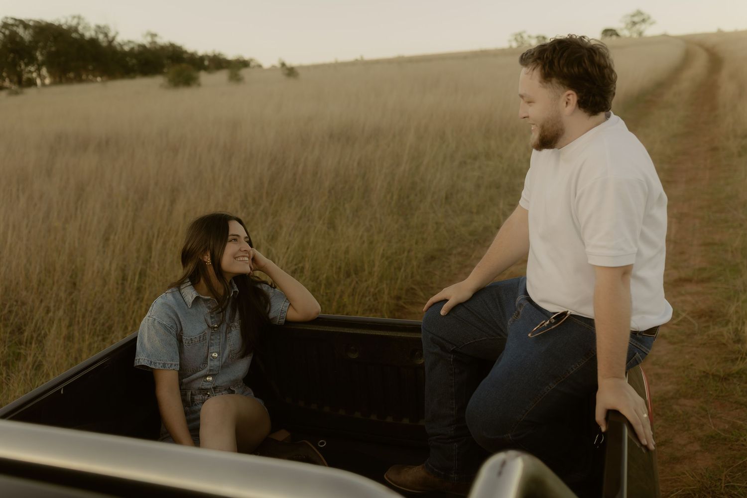 Couple sharing a candid moment at sunset, woman sits in truck bed while man stands beside her in grassy field.