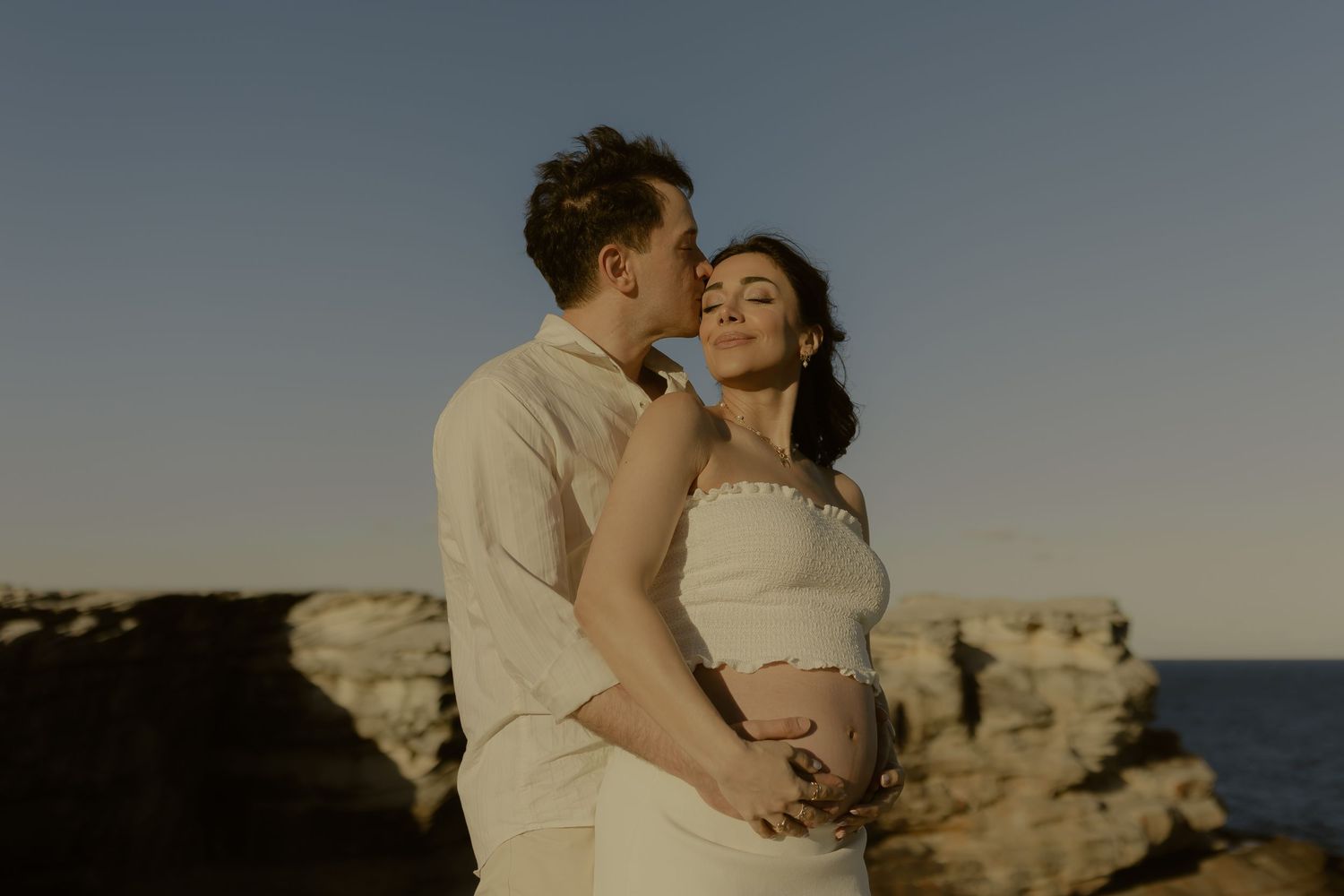 Couple embracing on rocky coastal cliffs at sunset, pregnant woman in white dress smiling serenely.