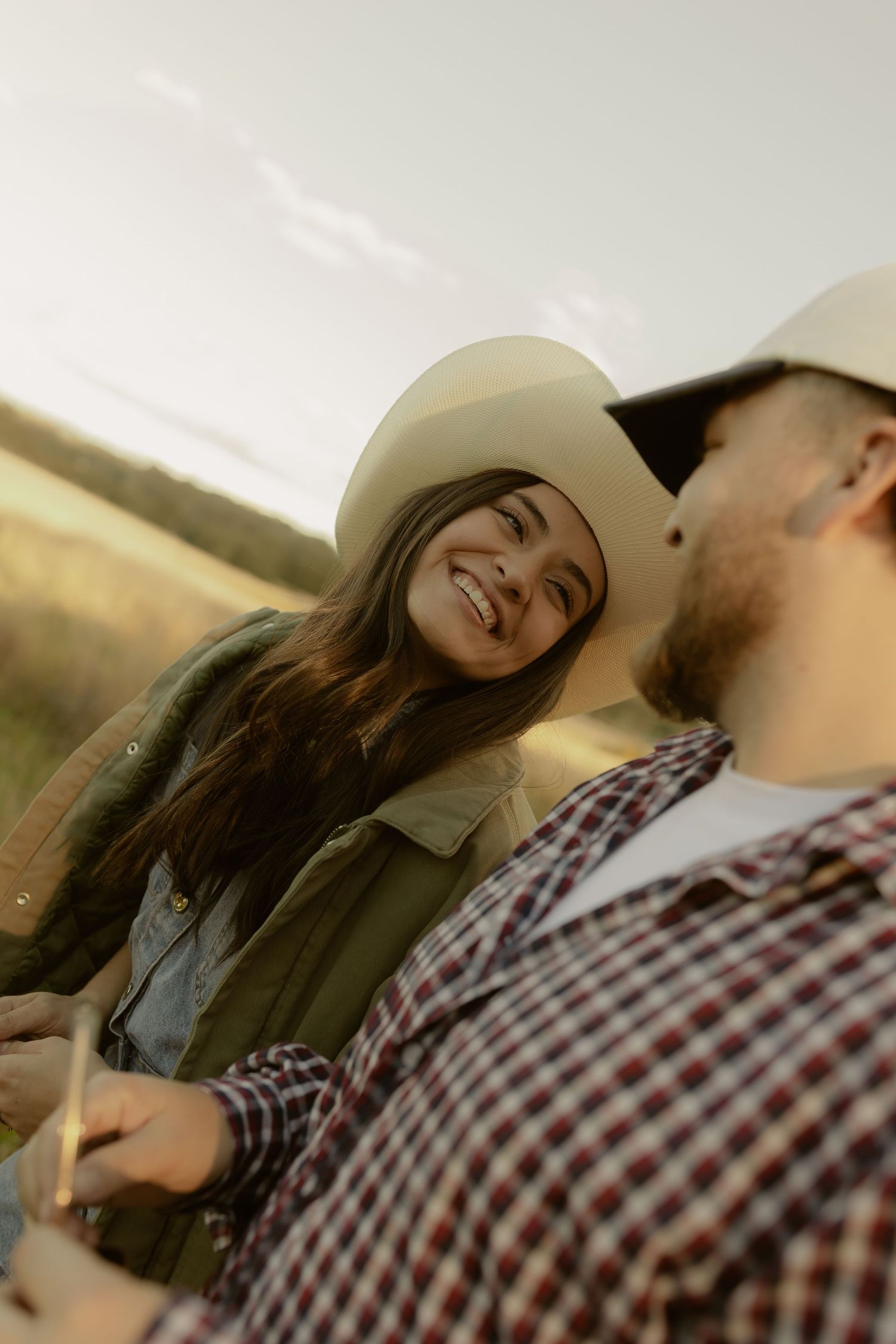 Smiling woman in a cowboy hat gazes at a man in a plaid shirt during a golden hour outdoor photo session.