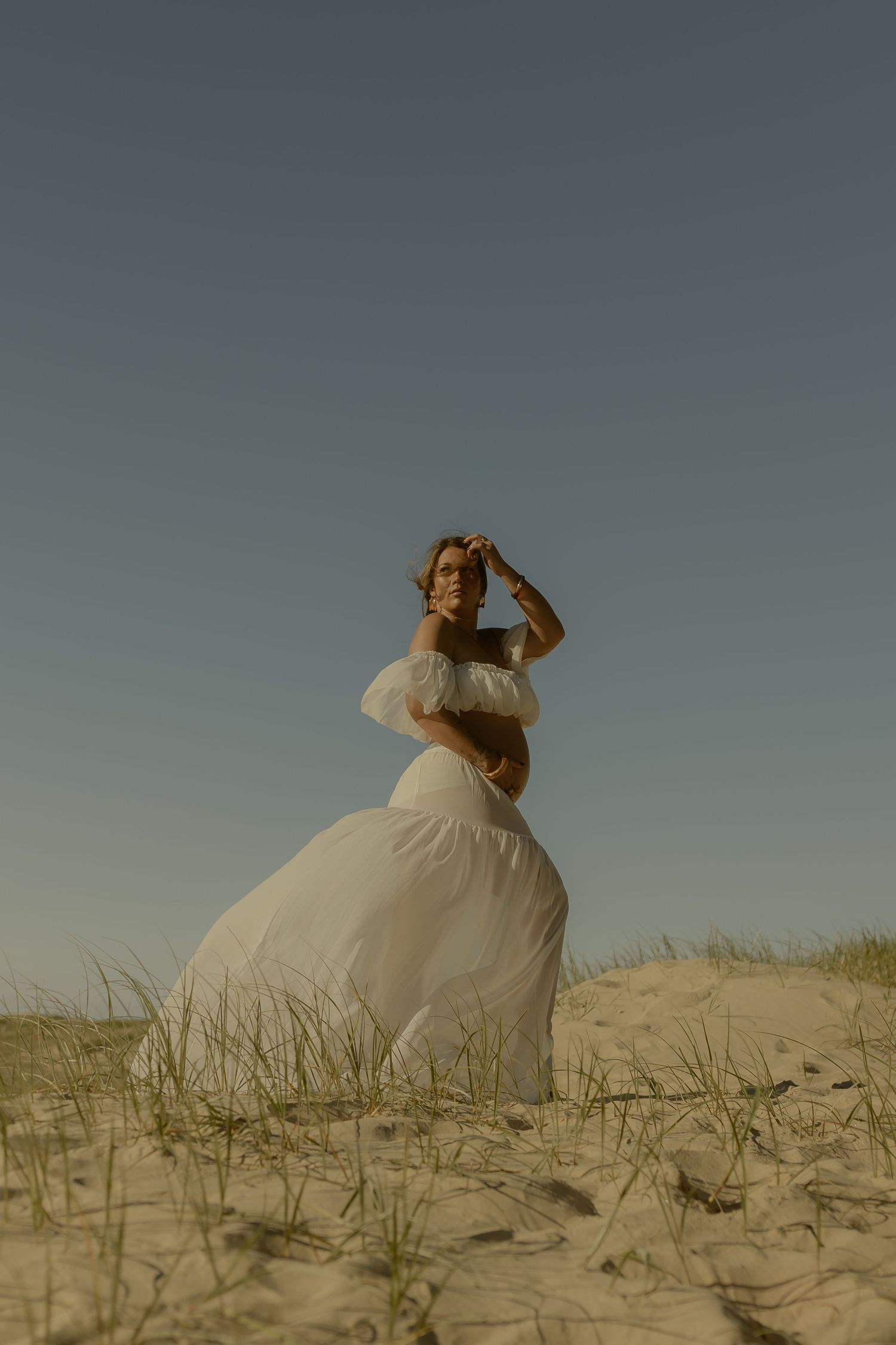 Pregnant woman in sheer white gown poses gracefully on sandy dunes, shielding eyes from sunlight.