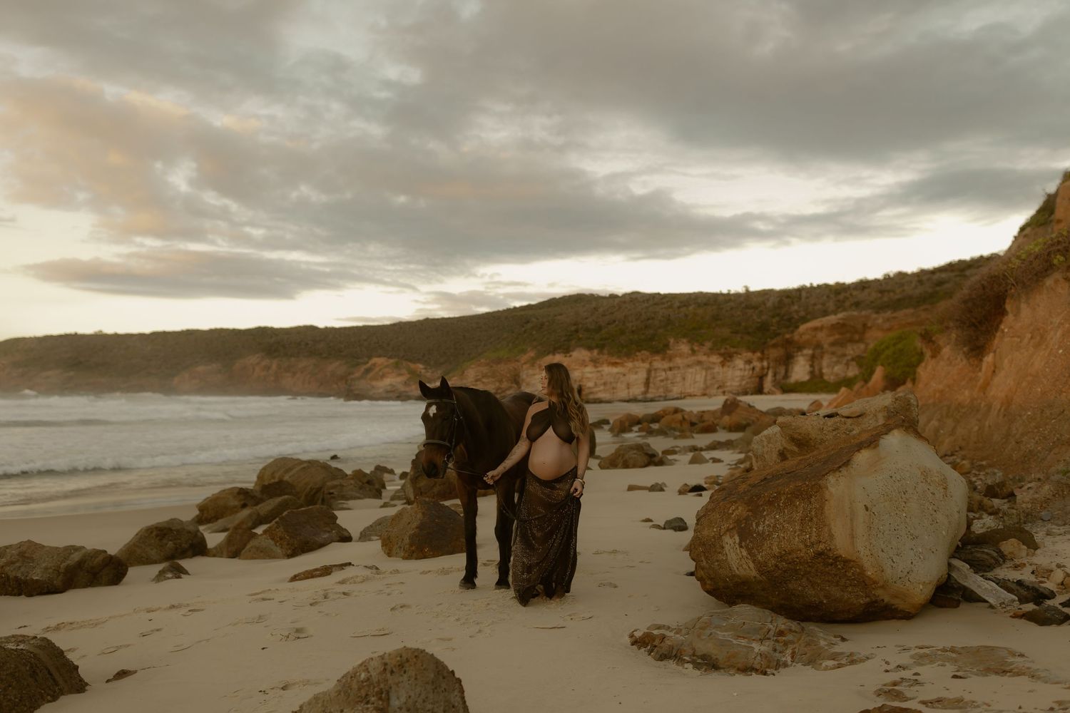 Pregnant woman in flowing dress stands with dark horse on rocky beach at sunset with dramatic cliffs.