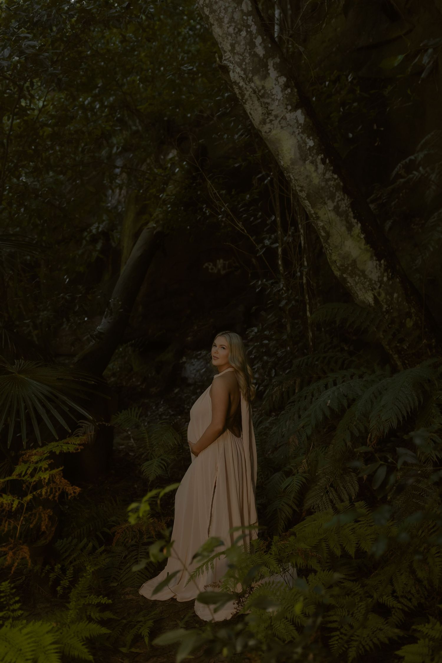 Pregnant woman in blush gown standing in lush green forest, cradling baby bump surrounded by ferns and trees.