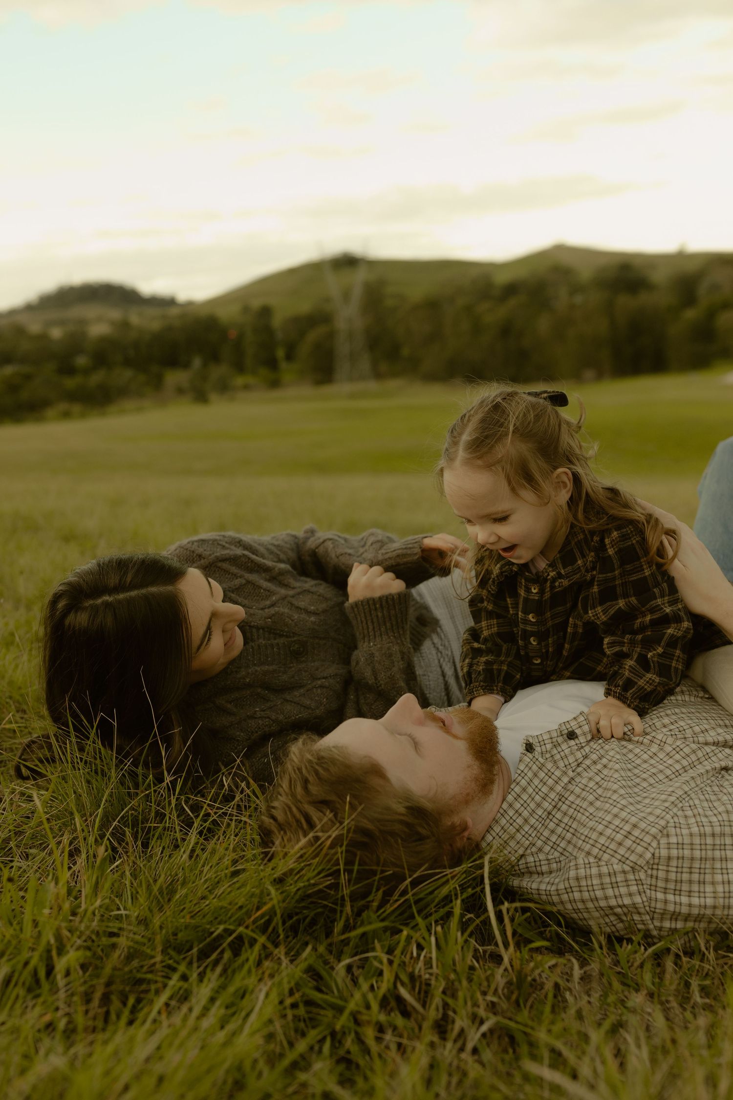 Family lying in grass at sunset, mother and daughter smiling at father.