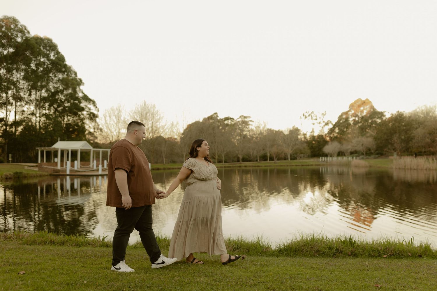 Pregnant couple holding hands walking by a serene lake at sunset in a lush green park.