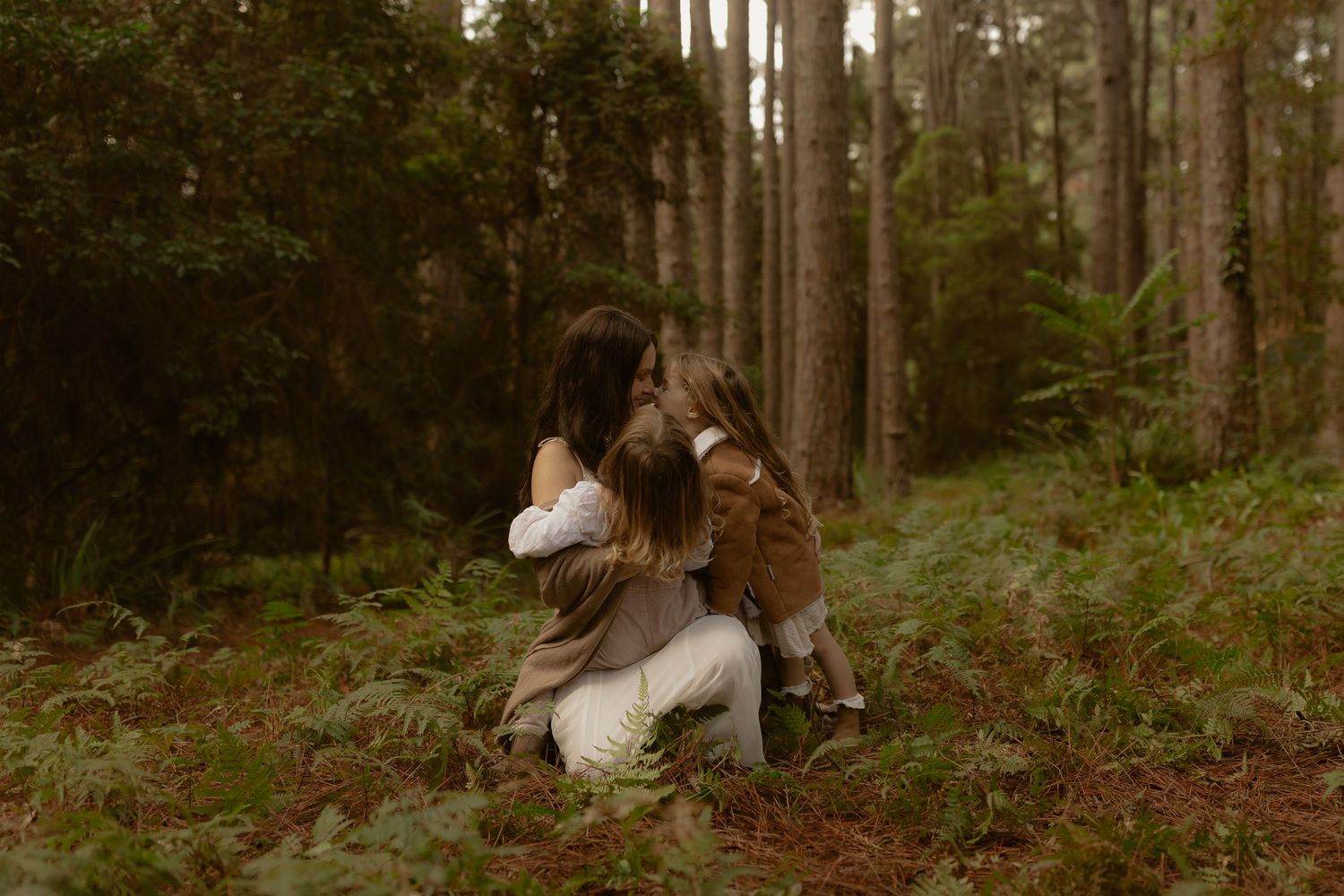 A mother kneels in a lush forest embracing her young child among ferns and tall trees.