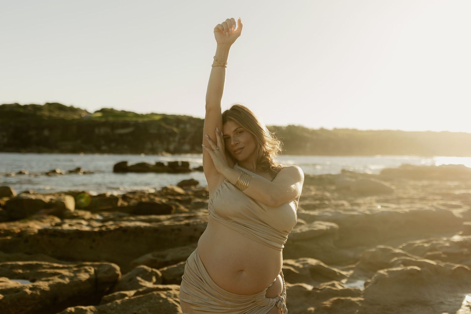 Pregnant woman posing on rocky beach at sunset with arm raised, wearing a beige crop top and skirt.