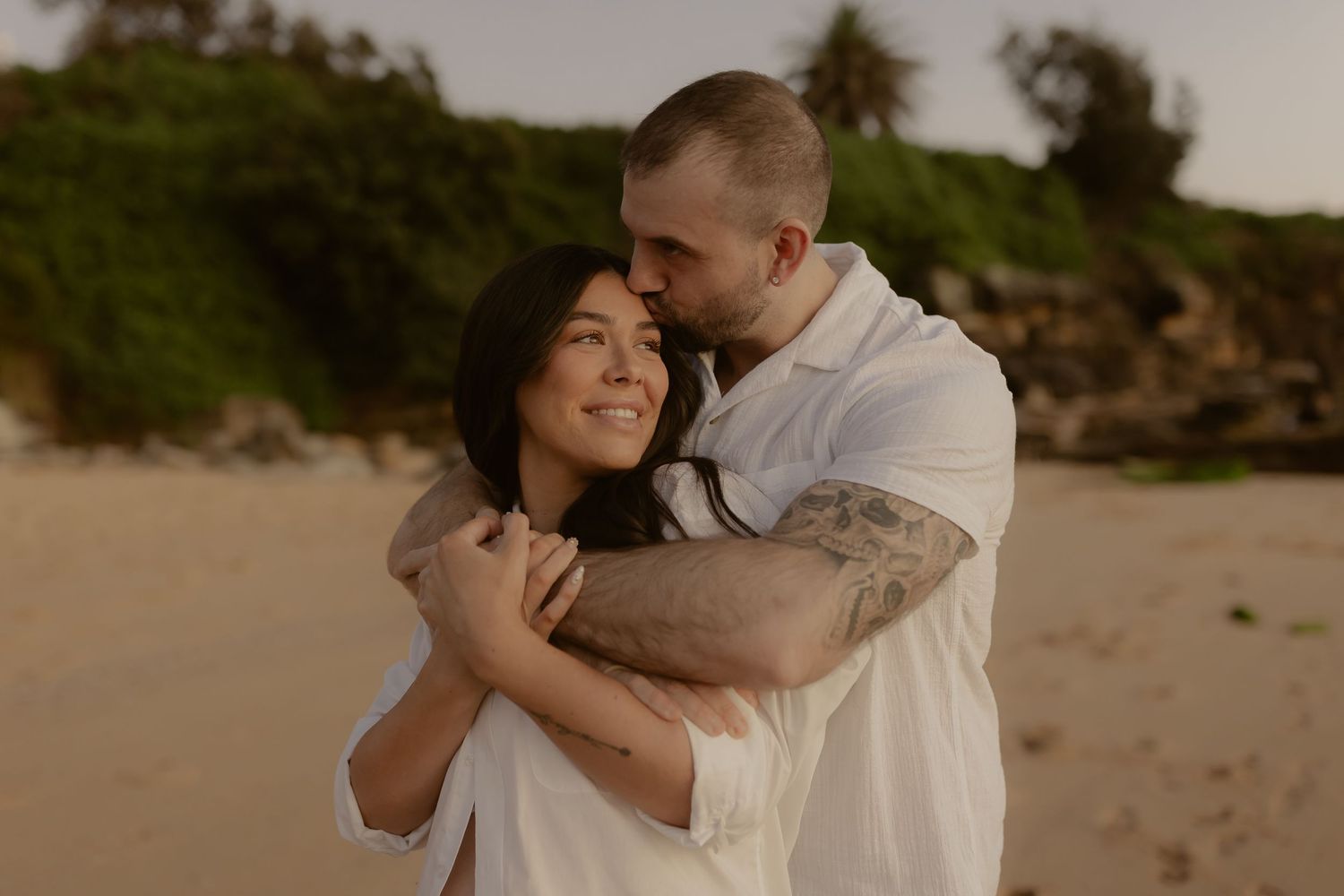 Couple embracing on sandy beach at sunset, man with tattoos kissing womans head.