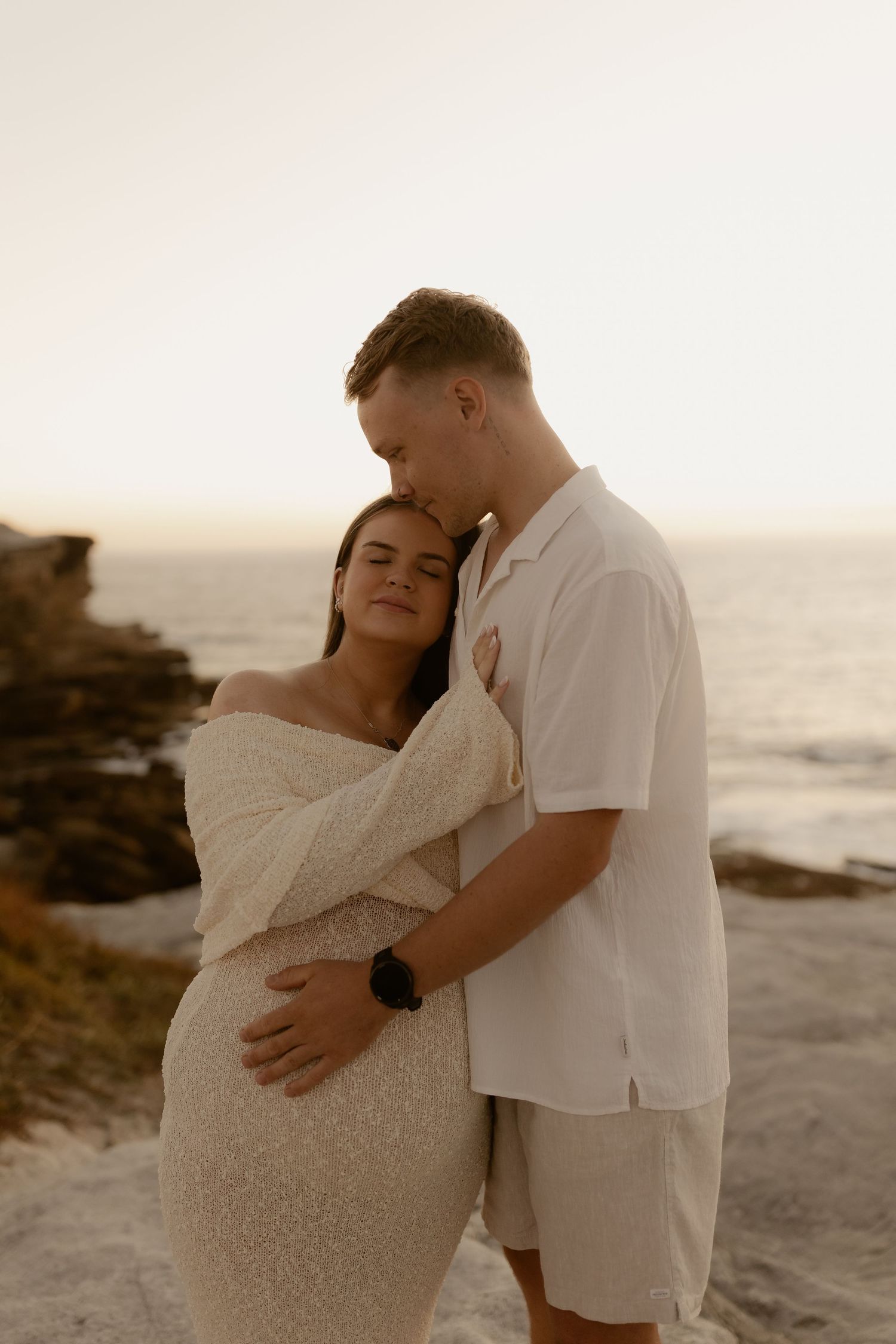 A couple shares intimate moments at sunset on a rocky beach in a series of romantic maternity photos.