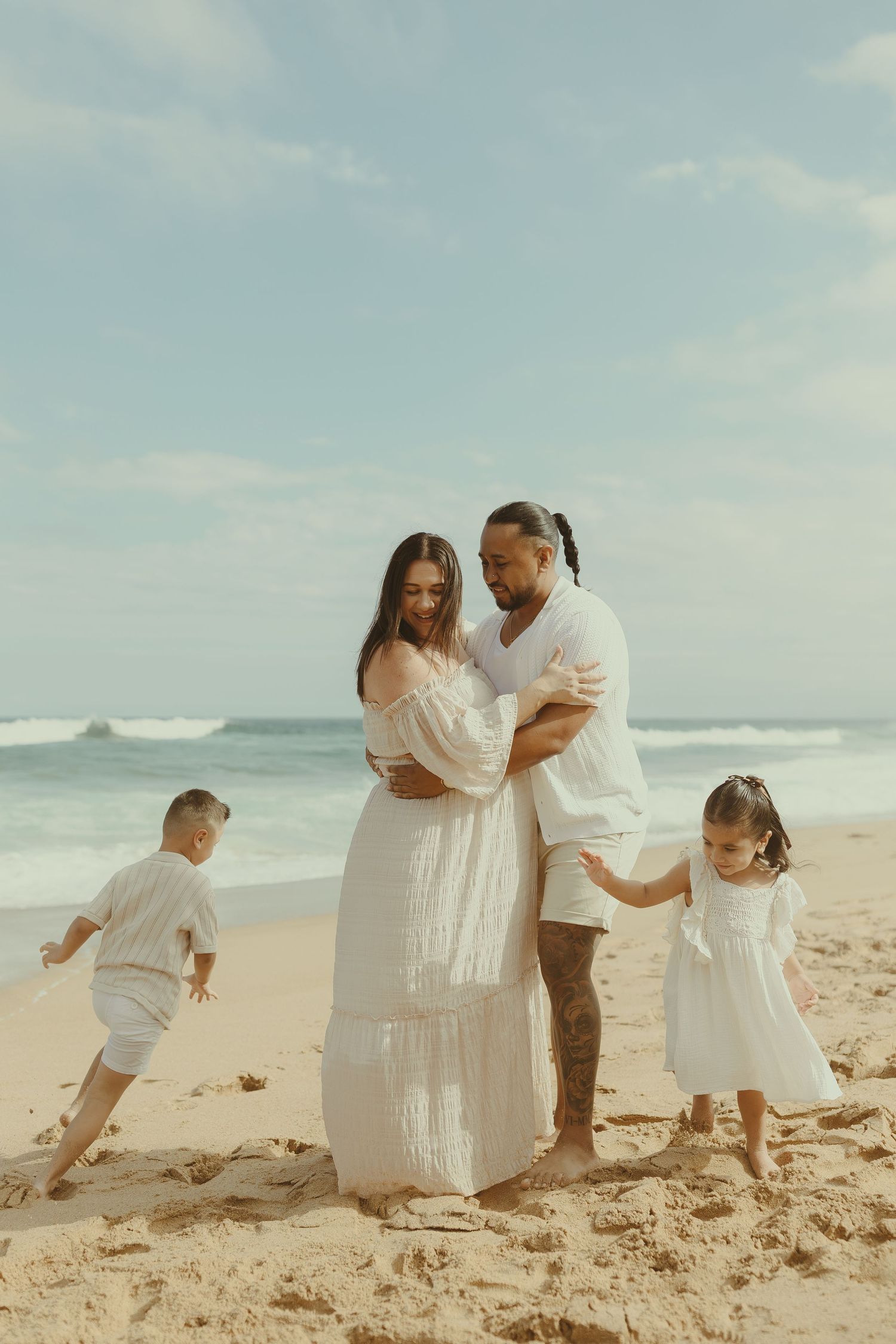 A family in white outfits plays together on a sandy beach with ocean waves in the background.