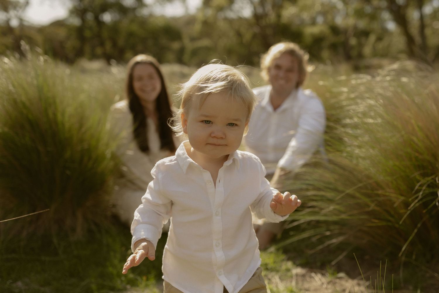 A group of people in white clothing walk through a sunlit field with tall grass and natural vegetation.