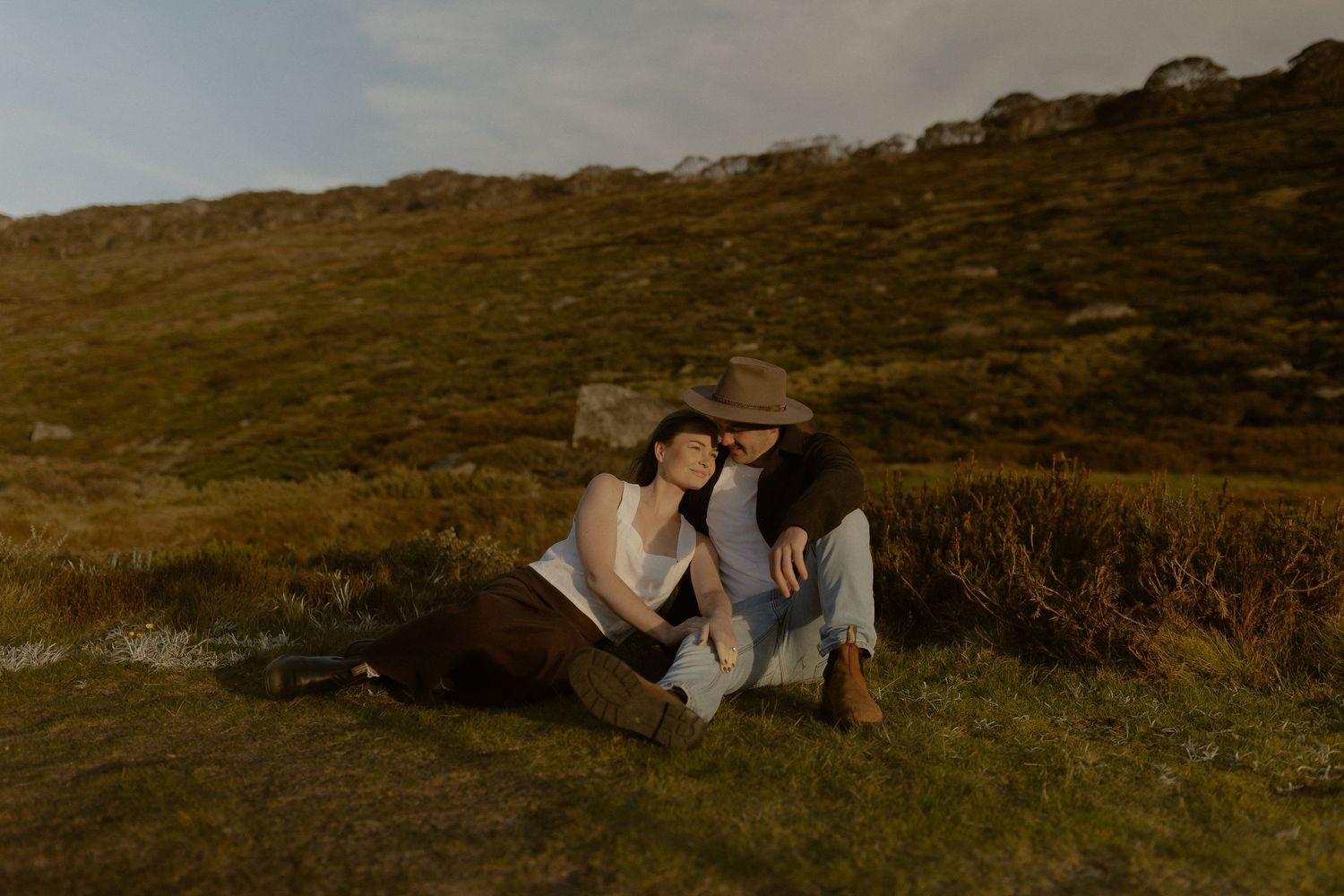 Two people share an intimate moment while sitting together on a grassy hillside at dusk.