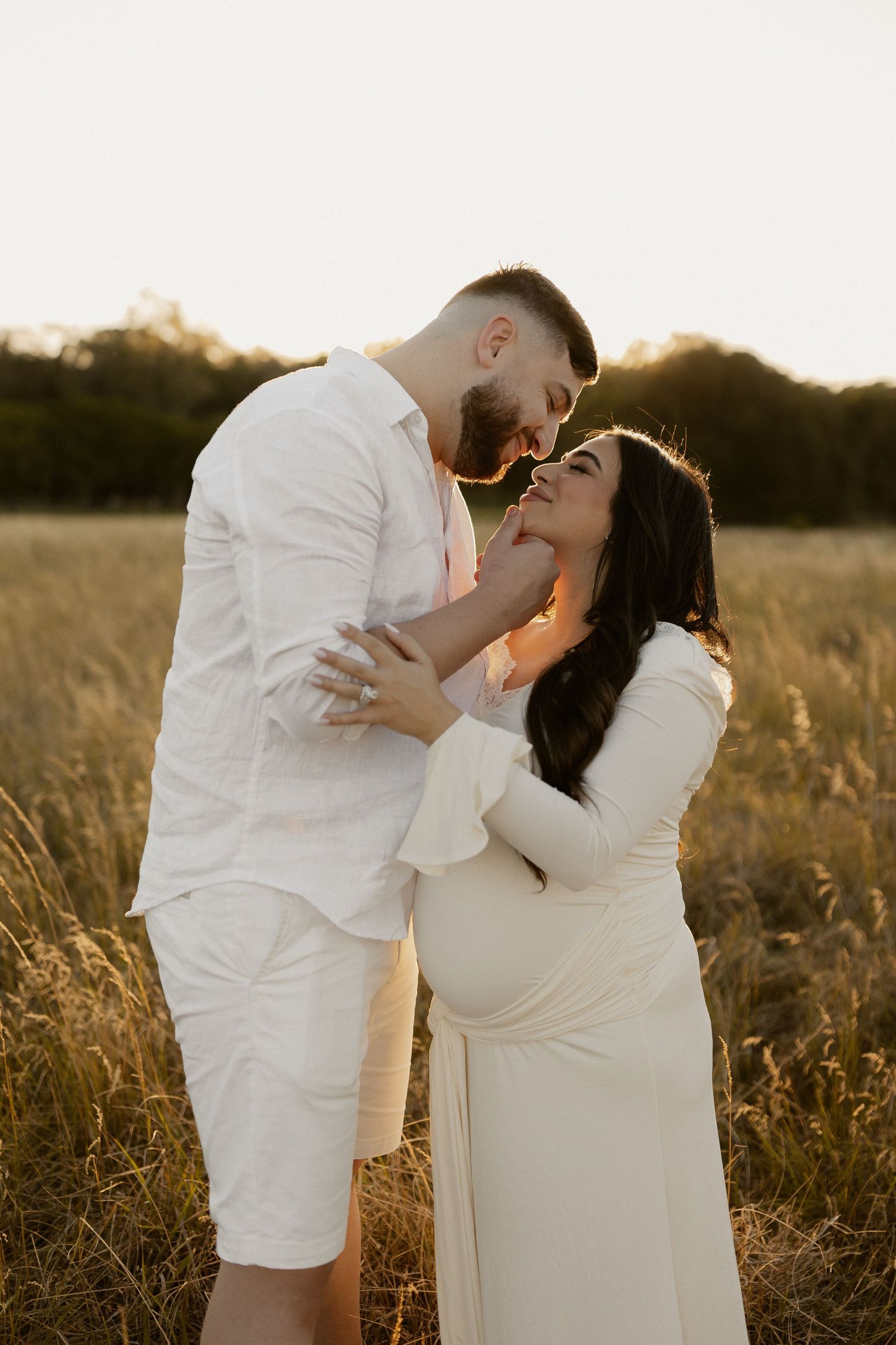 Intimate maternity portrait of couple in white clothing laughing together in sunlit meadow.