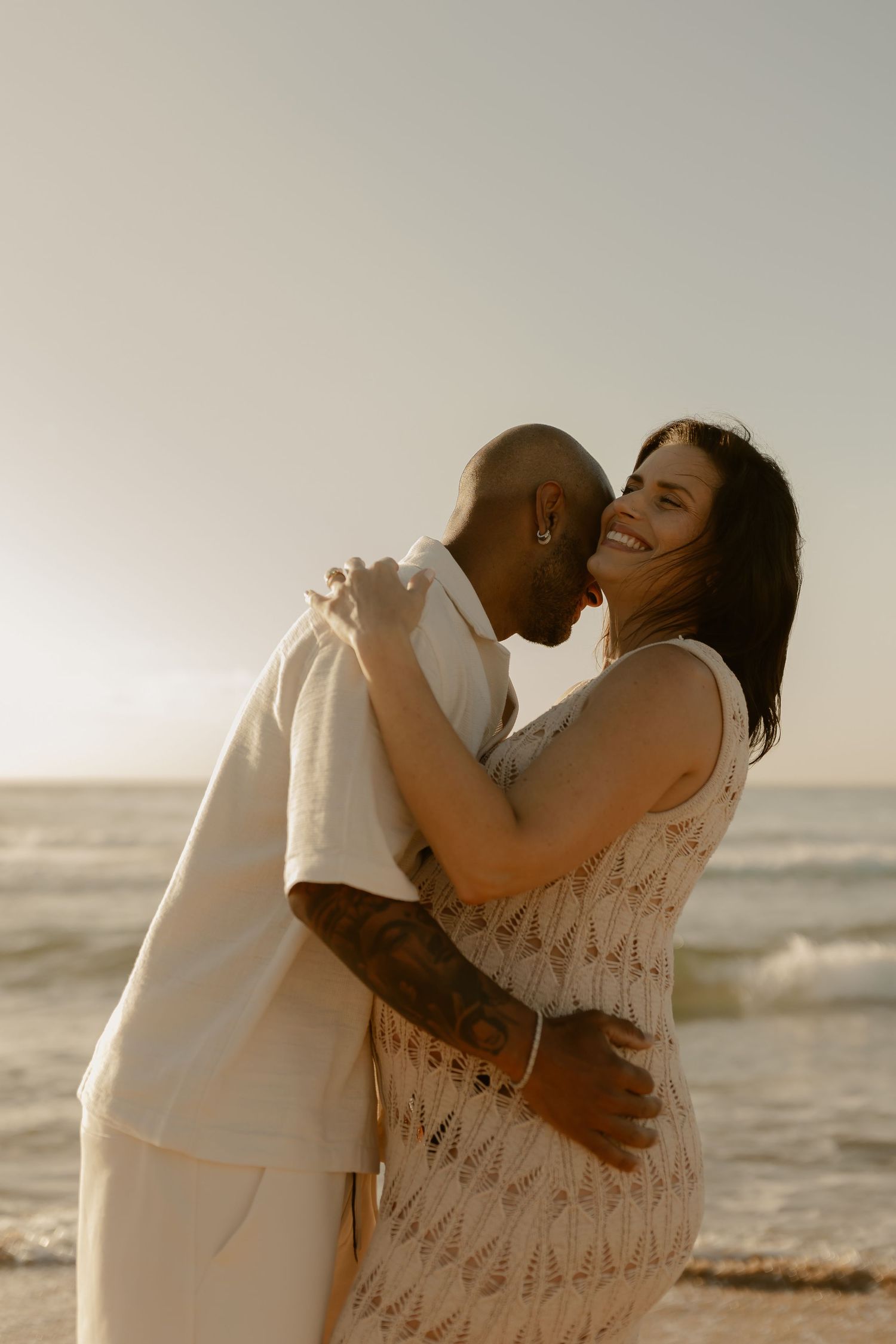 A couple embraces and laughs together on the beach during sunset.