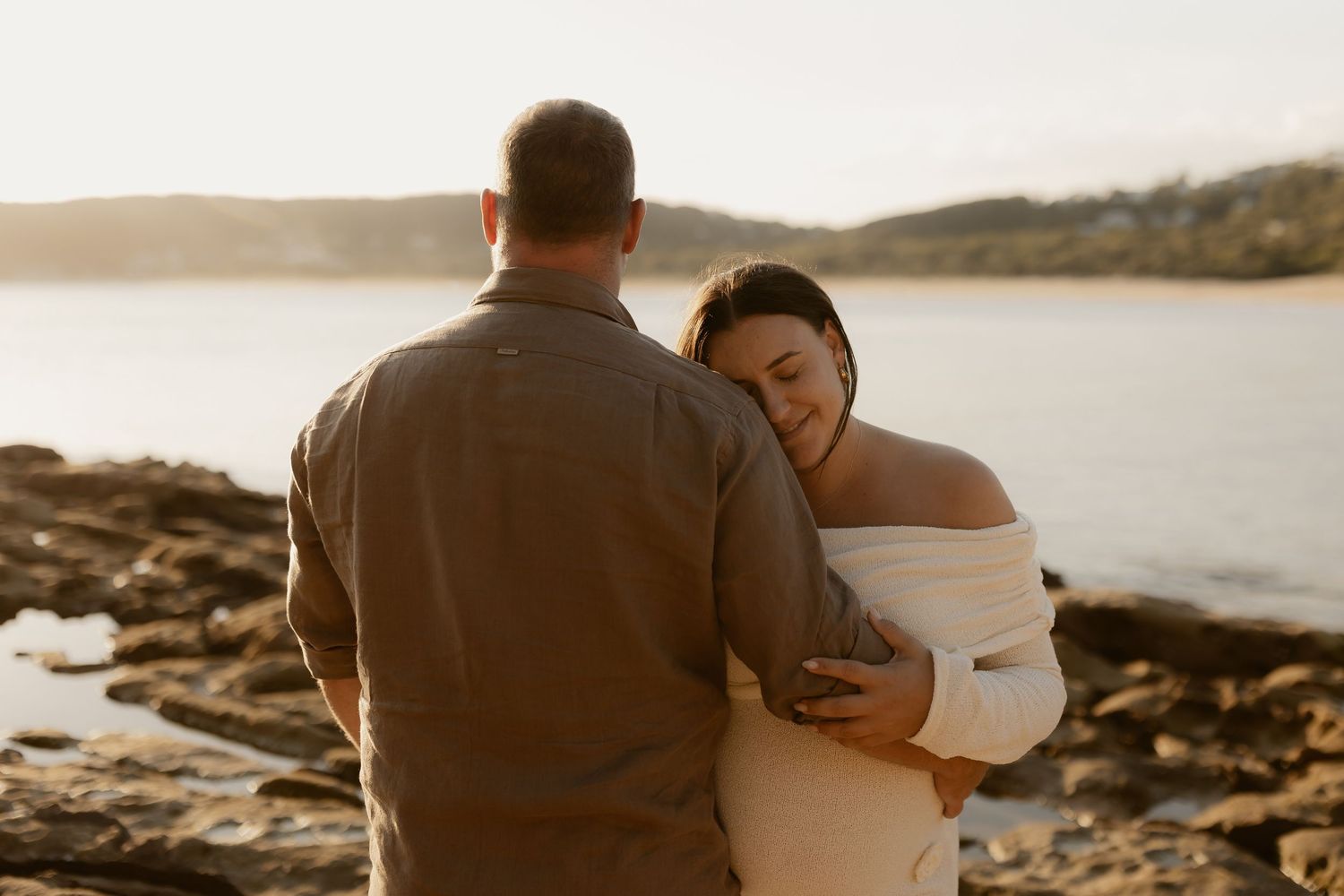 Silhouetted couple embraces while watching sunset over rocky coastline.
