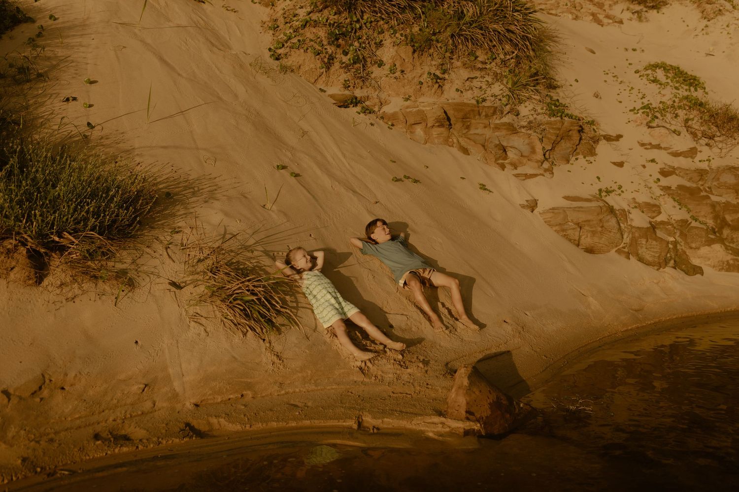 Kids slide down a steep sandy slope at the beach during golden hour.