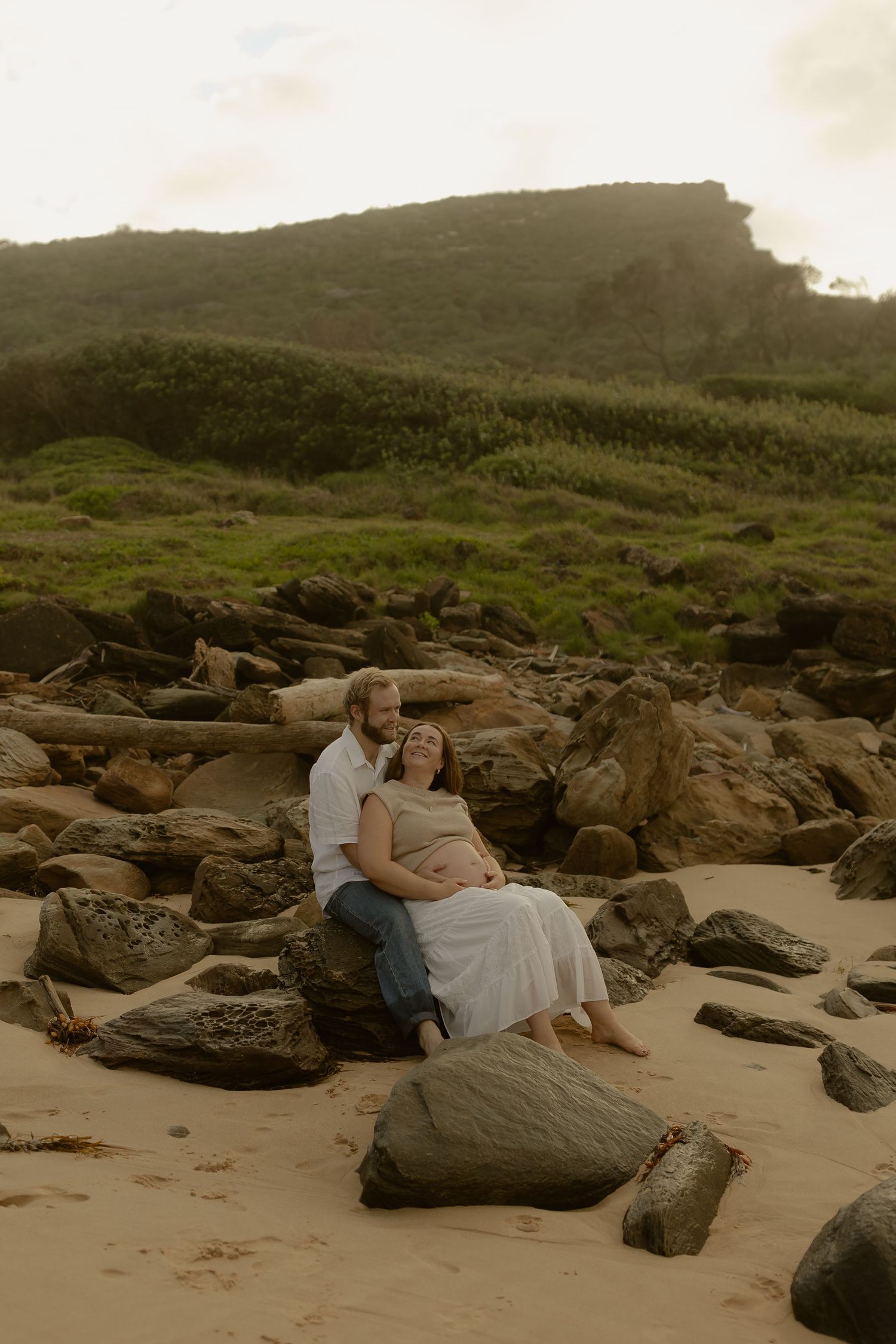 A couple sits together on large rocks at a scenic beach during golden hour.