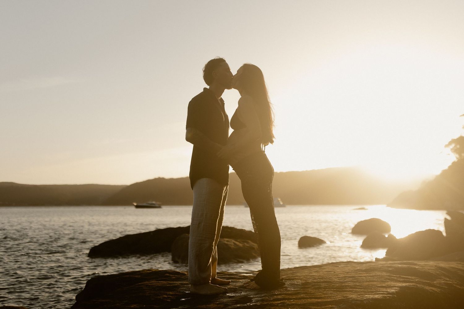 Silhouettes of couple embracing at sunset on a rocky beach with golden sunlight in background.