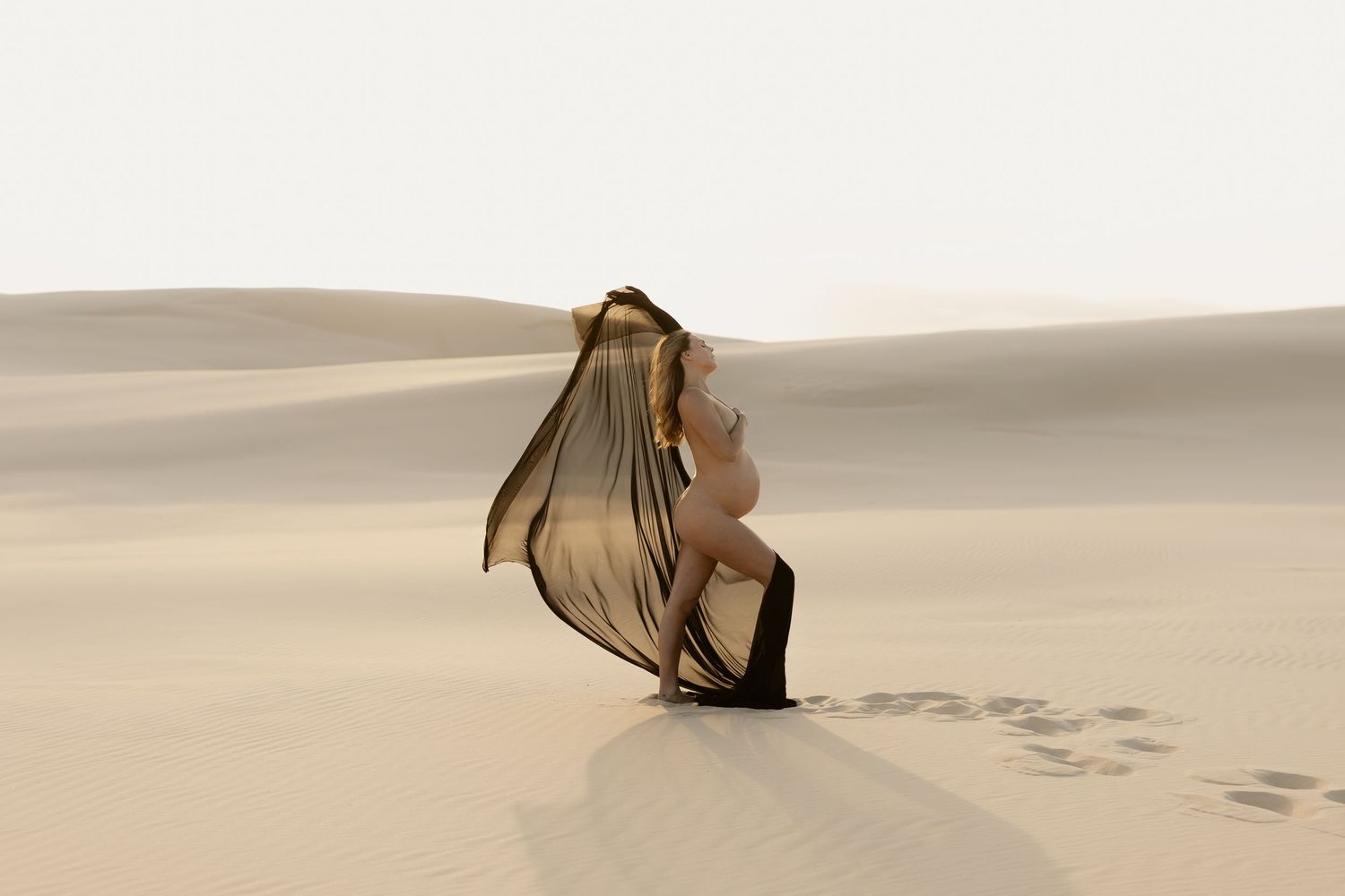 Figure in flowing dress performs elegant dance movements across pristine desert sand dunes at sunset.