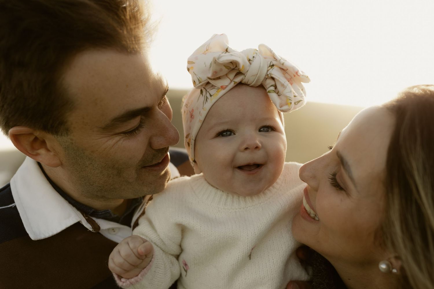 Happy baby in floral headband smiles between two loving parents in warm golden sunlight outdoor family portrait.