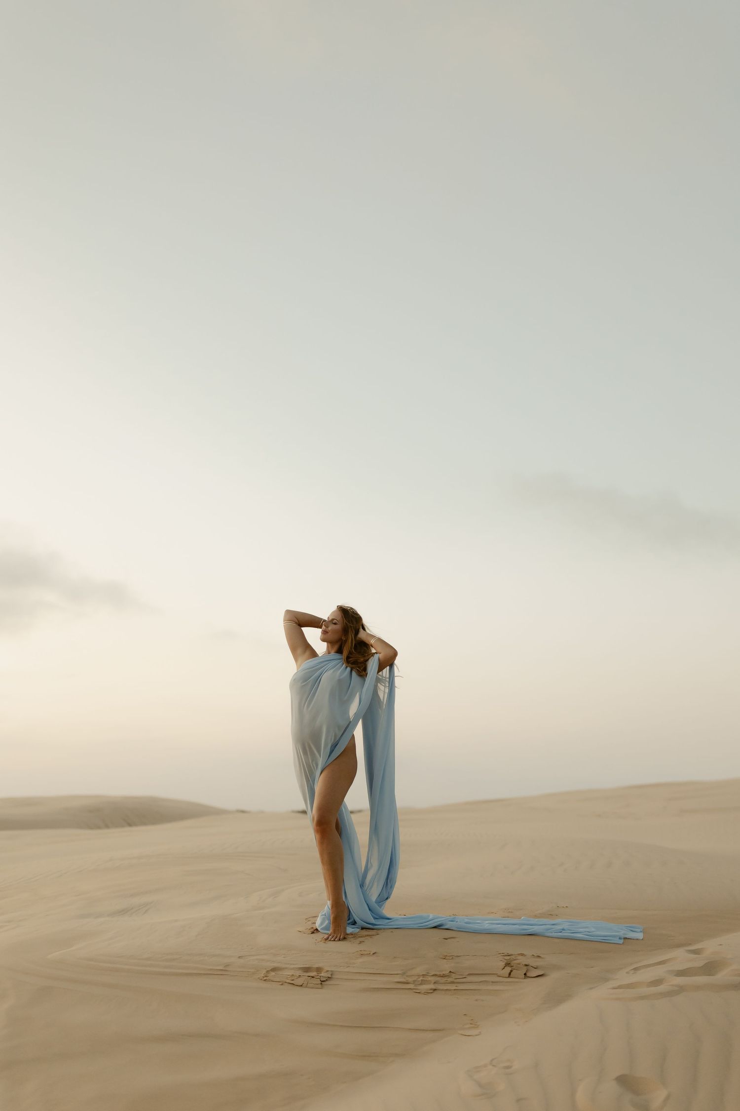 A person in flowing blue fabric dances gracefully across desert sand dunes at sunset.