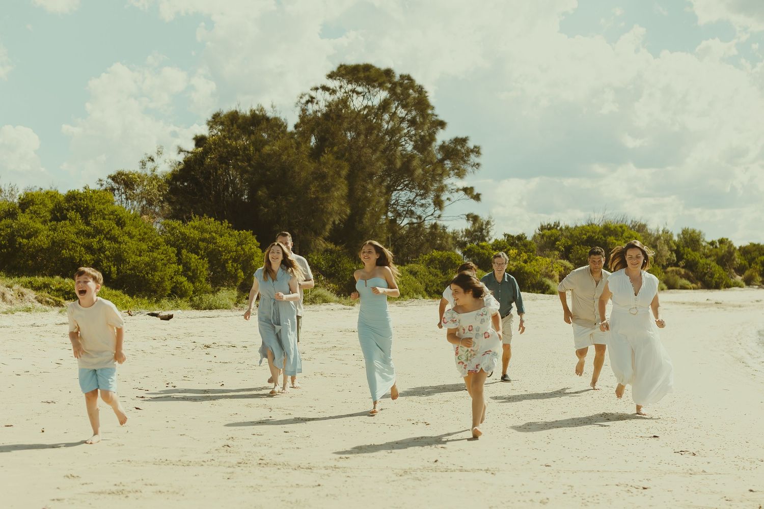 Group of people joyfully running together on a sunny beach with trees in the background.