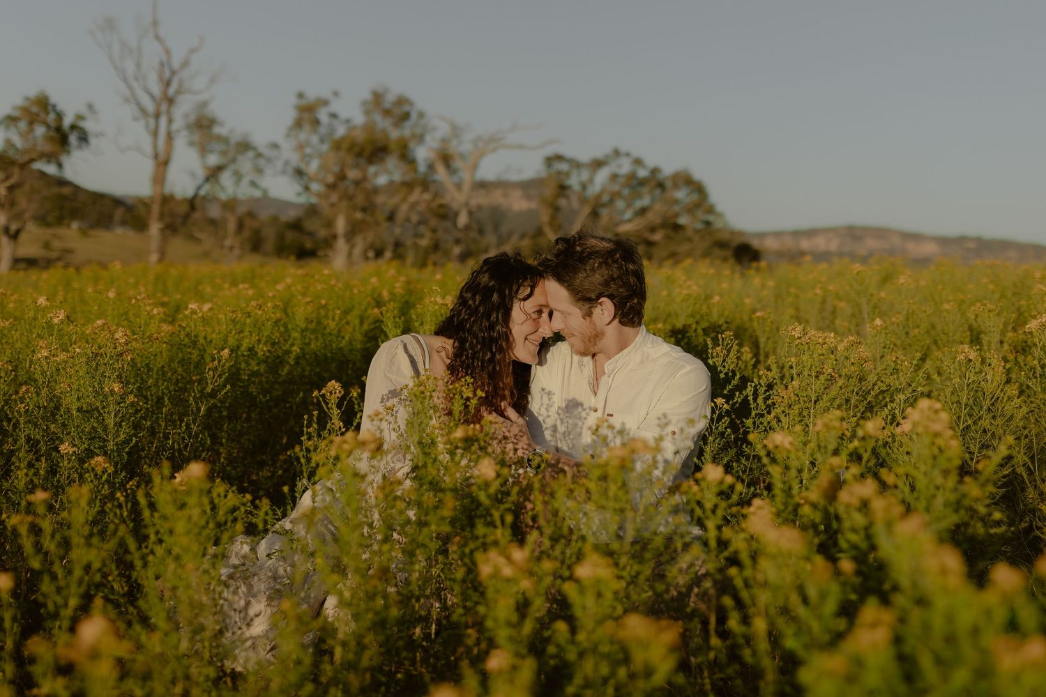 Couple embracing in distance amid tall wildflowers with mountains in background at sunset.