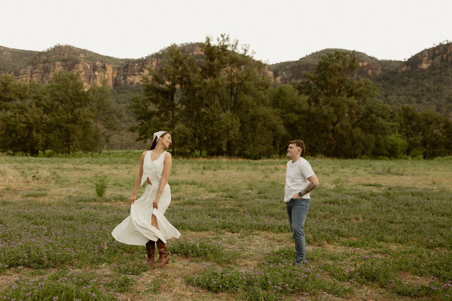 Two people walk playfully through a grassy field with mountains in the background at sunset.