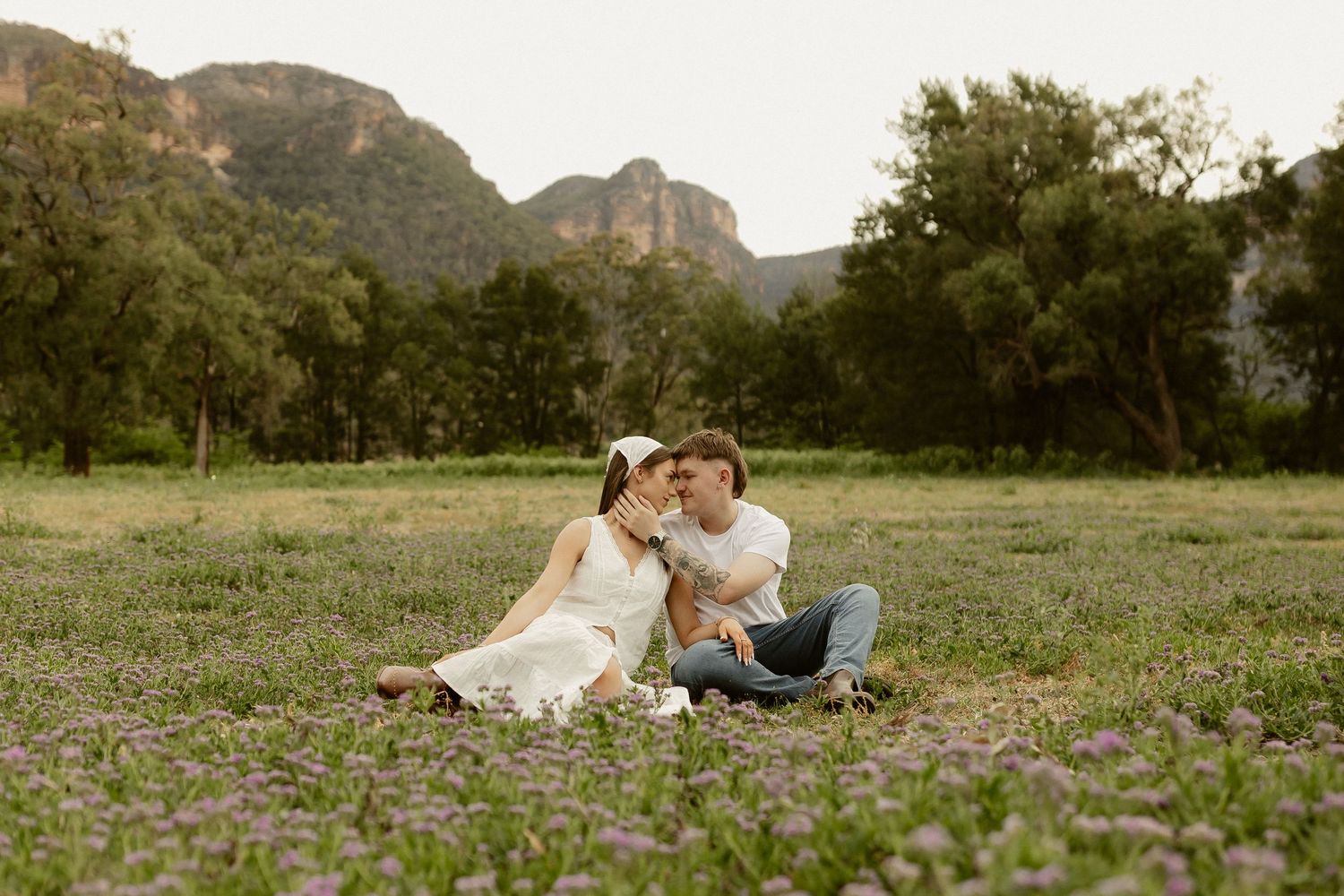 A couple sits together on wildflowers in a meadow with dramatic mountain cliffs in the background.