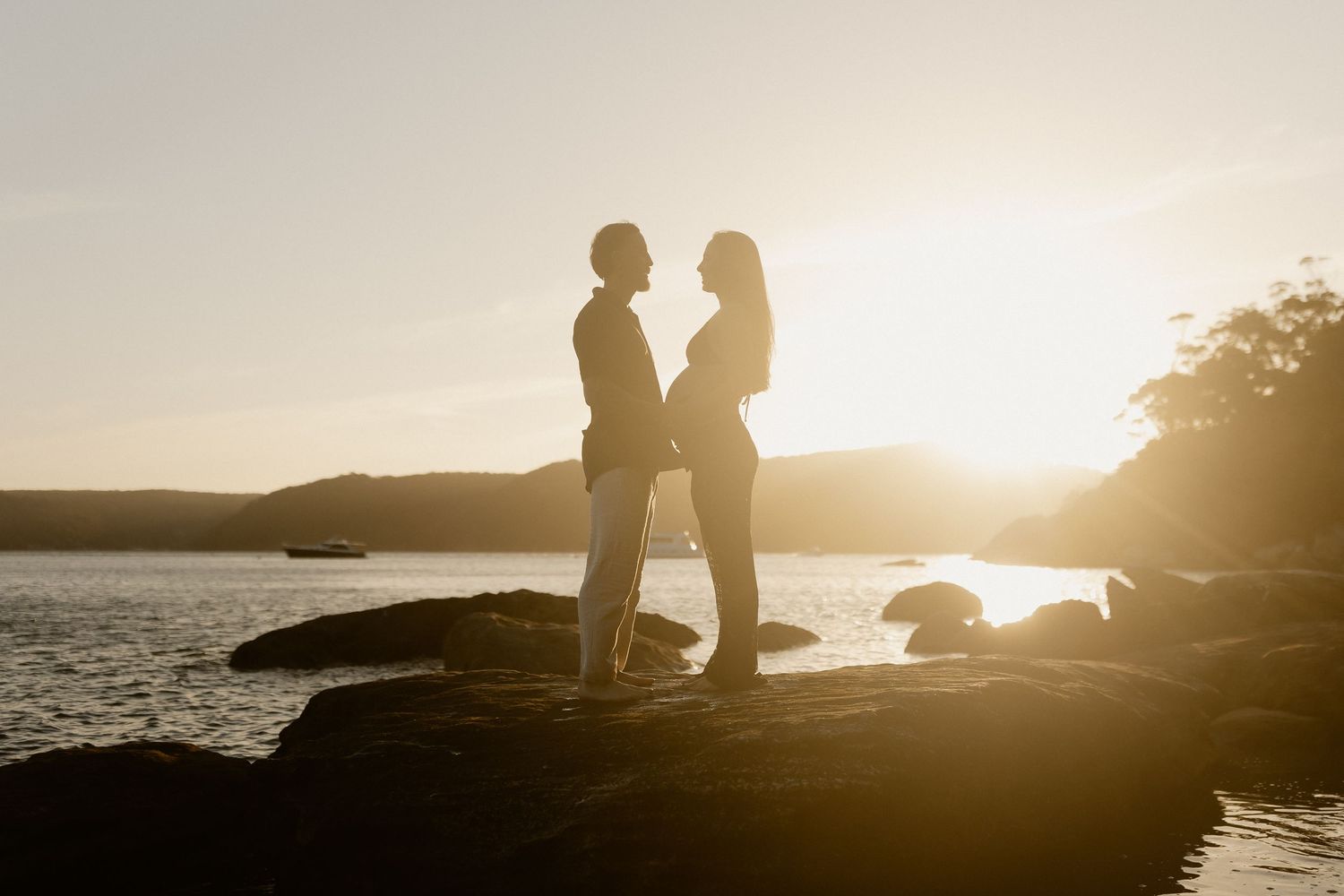 Silhouetted figures stand on rocky coastline during golden sunset over ocean waters.