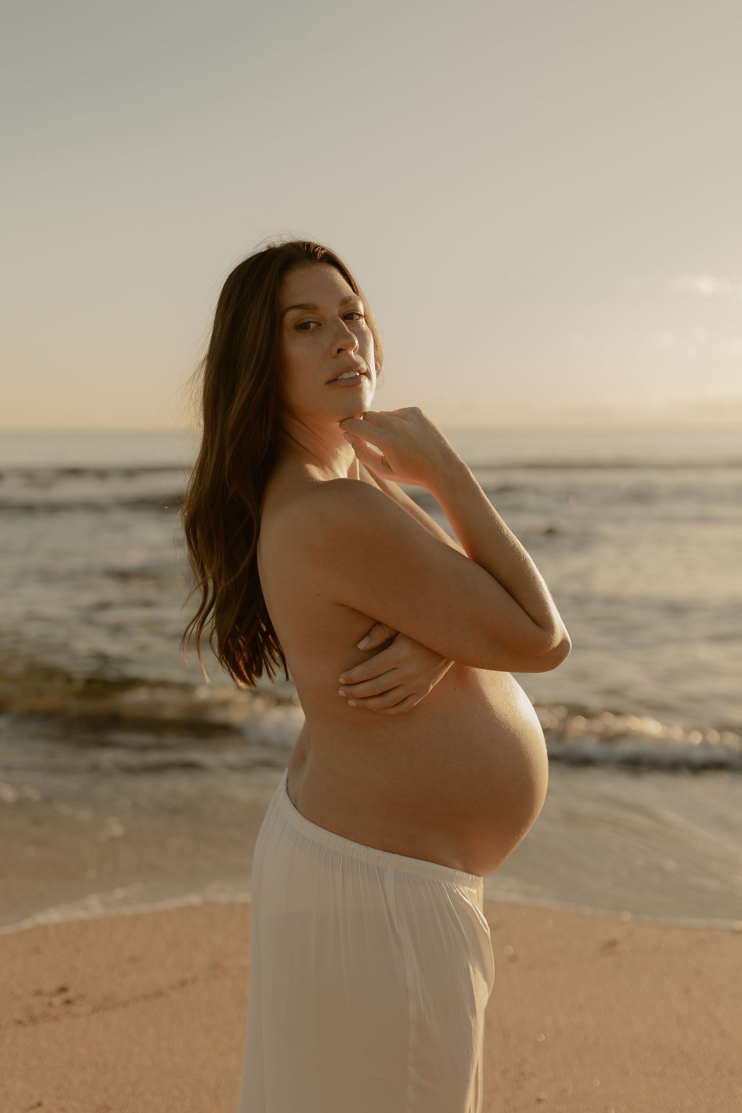 Maternity photoshoot on beach at sunset with woman in flowing white dress.