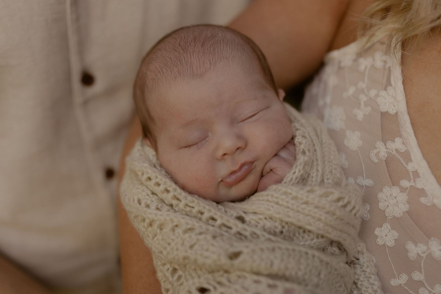 Peaceful newborn baby swaddled in cream knit wrap, sleeping in mother's arms during professional newborn photography session.