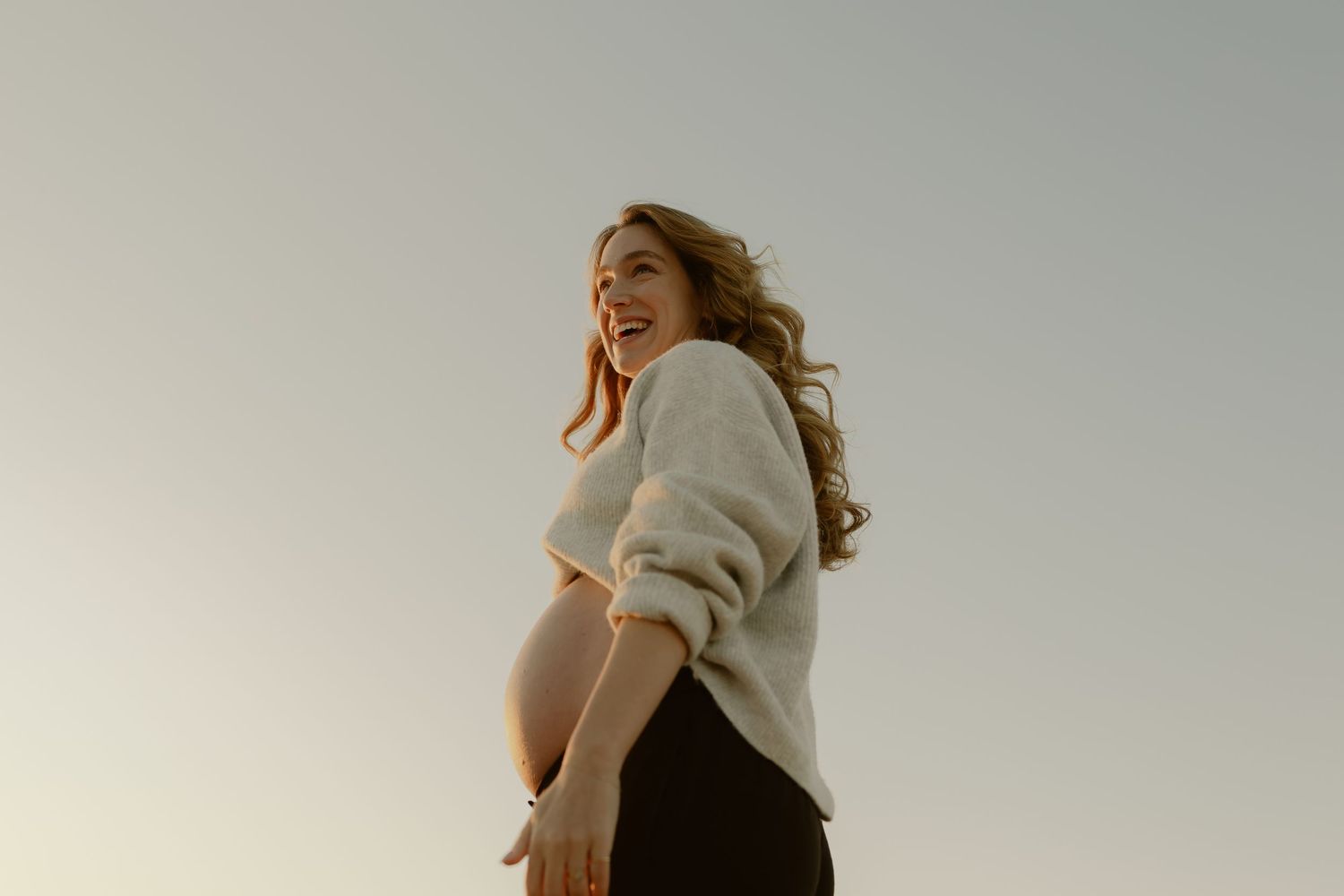 Happy pregnant woman in white sweater laughing outdoors against clear sky at sunset.