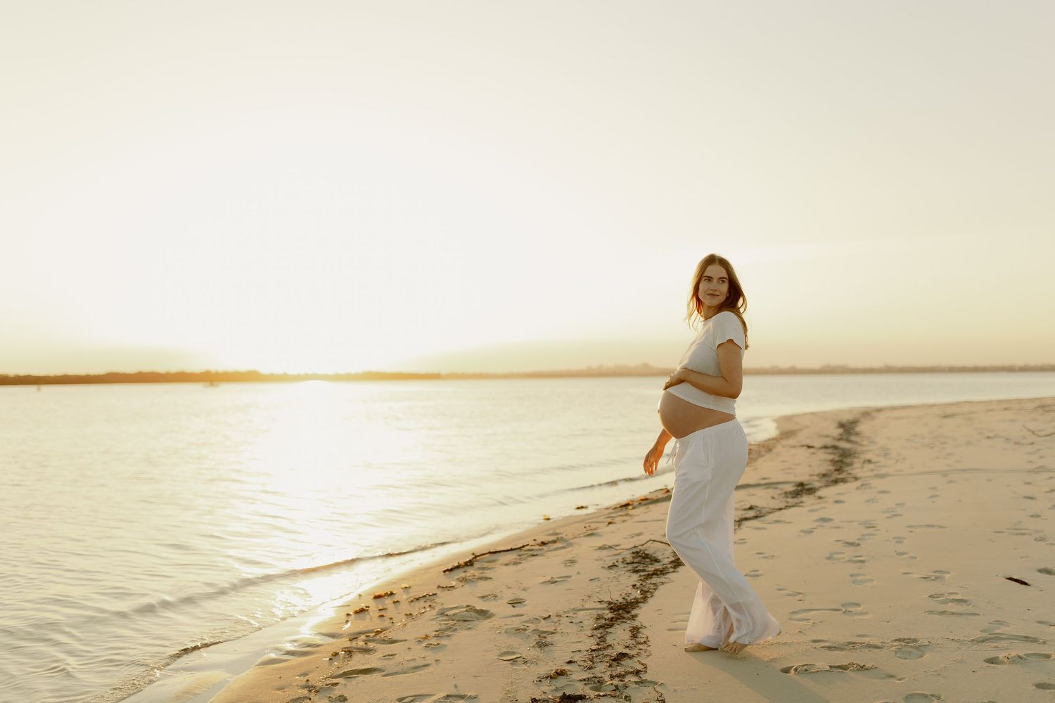 Pregnant woman in white outfit smiling on sandy beach at golden sunset by calm water.