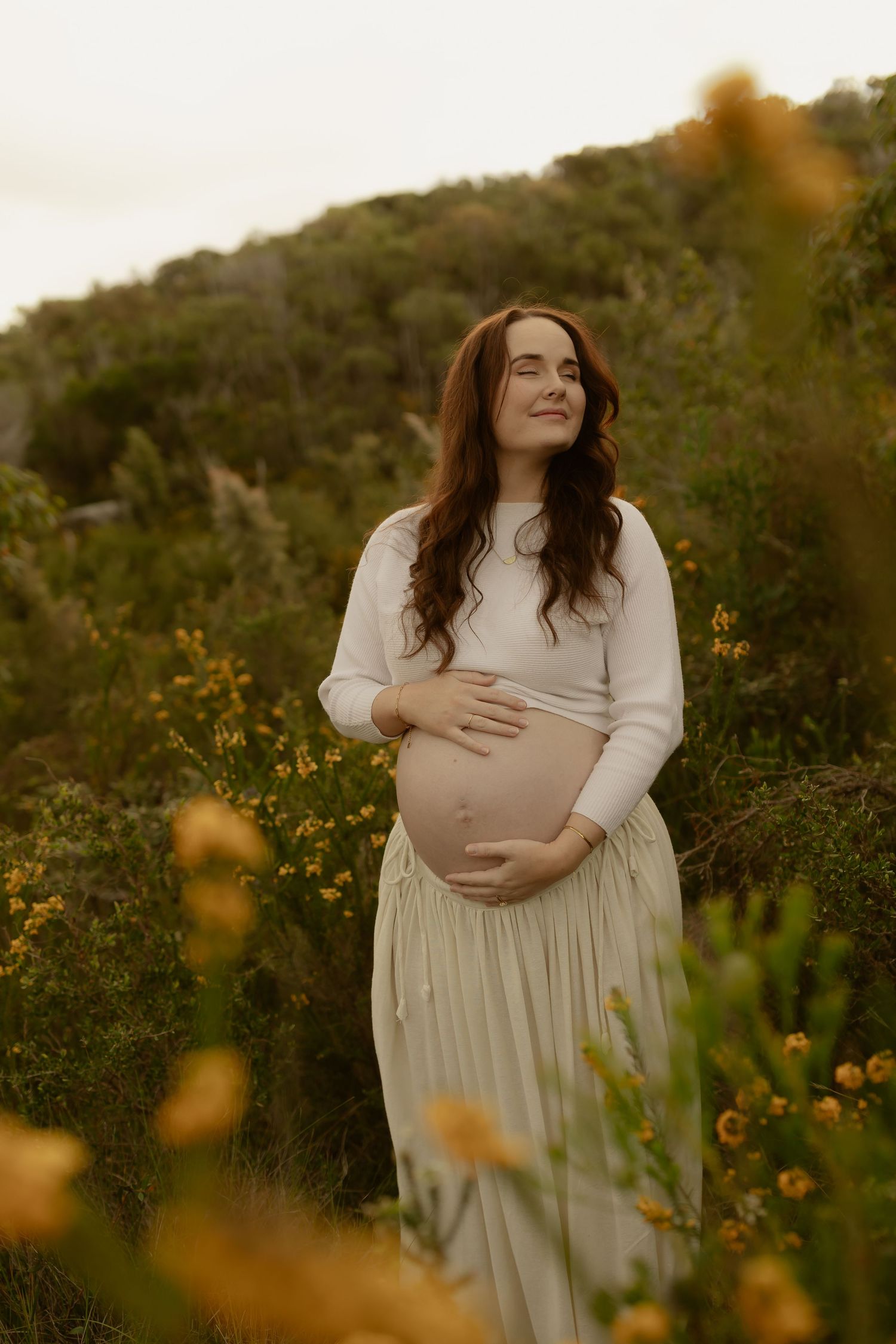 Pregnant woman in white crop top and cream skirt cradling her baby bump outdoors among yellow wildflowers.