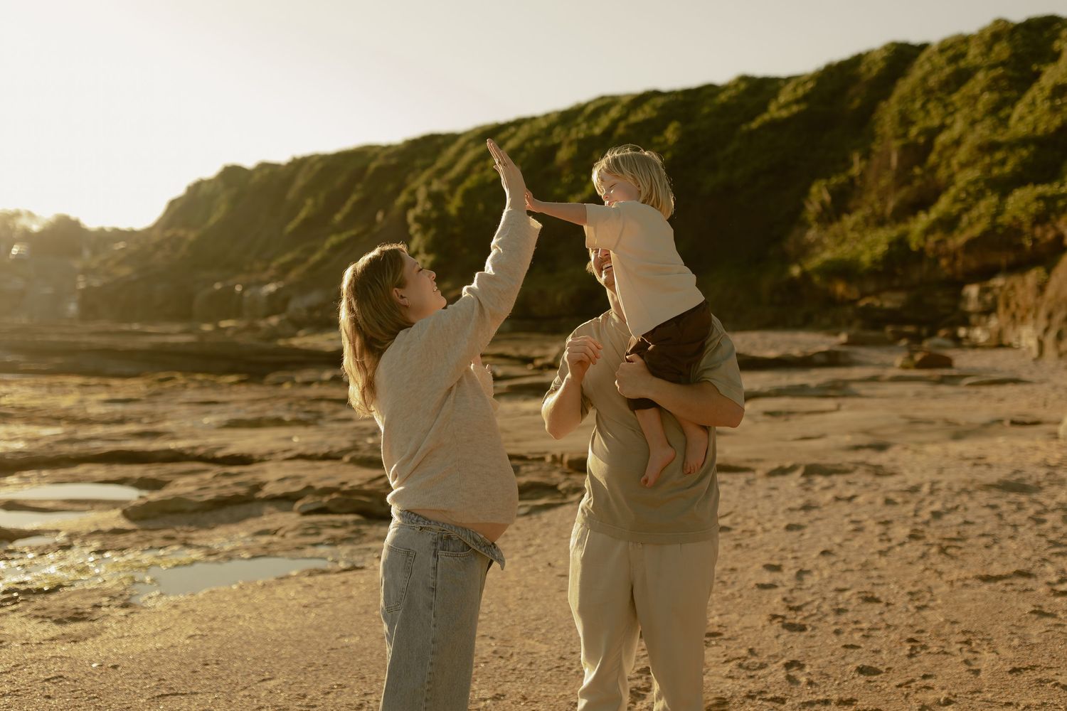 Parents playfully lifting their blonde toddler on a scenic sunlit sandy beach.