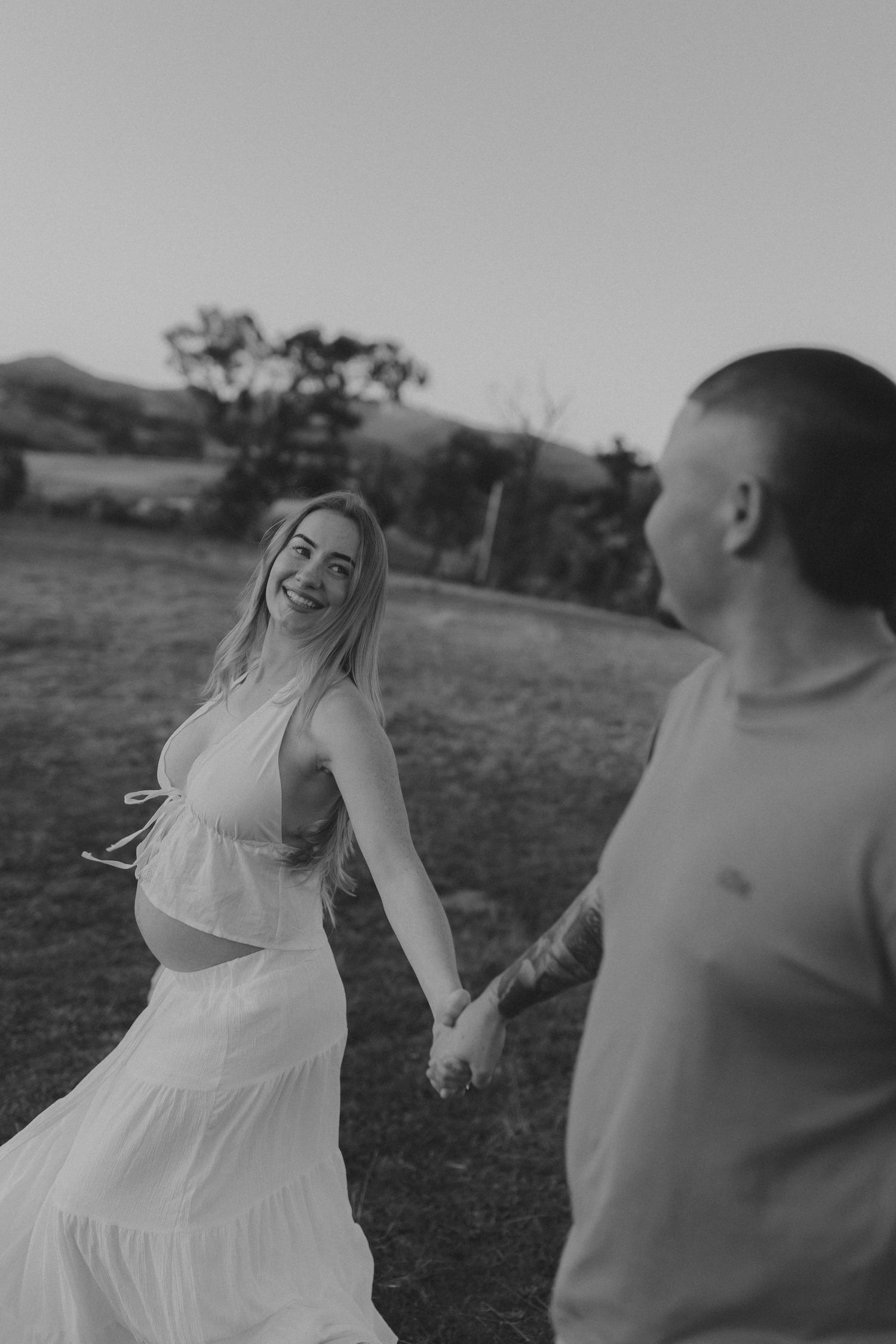 Black and white photo of smiling pregnant woman holding hands with partner in field.