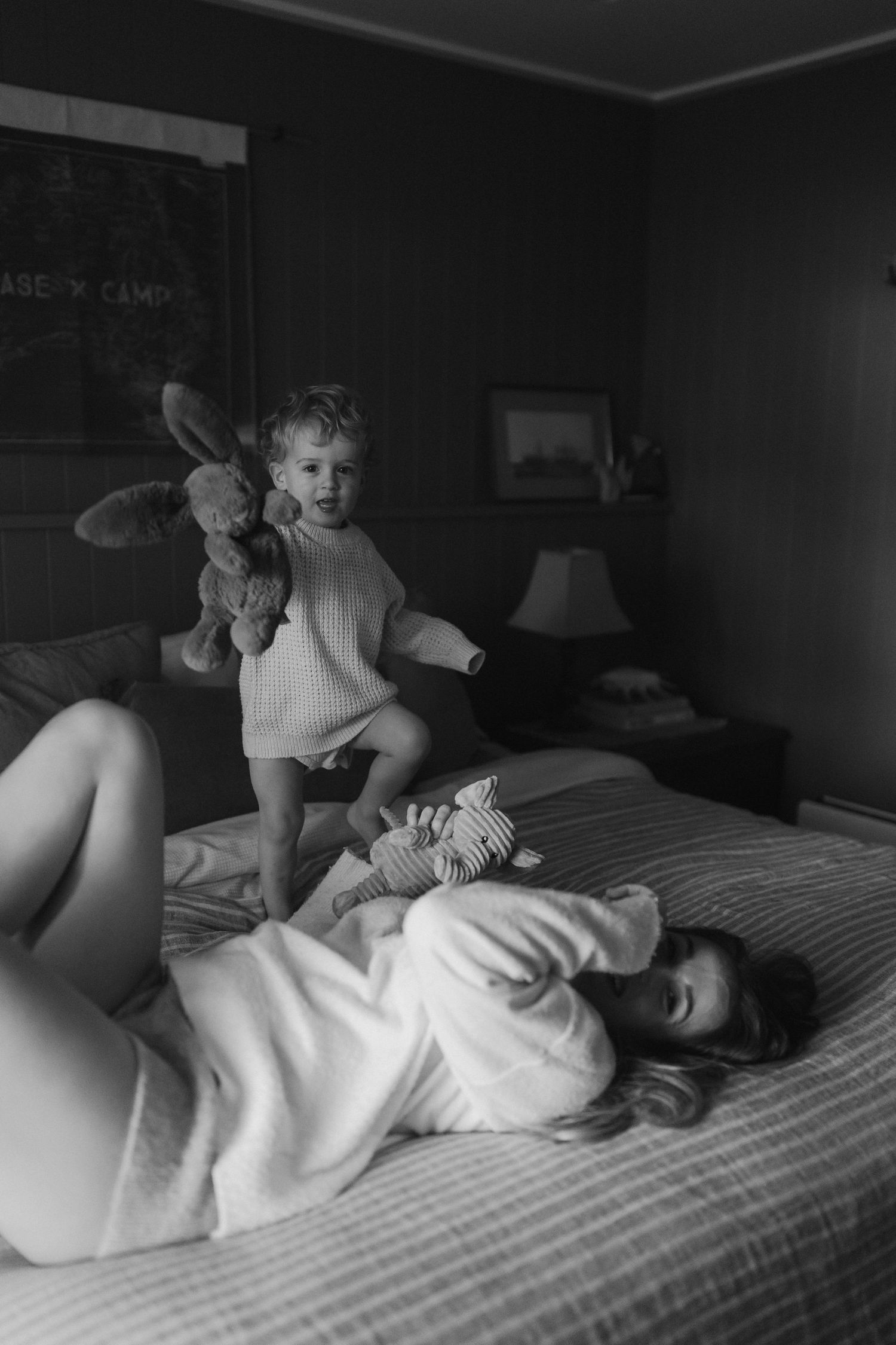A playful moment captured in black and white as a toddler in a sweater jumps on a bed with a stuffed toy.