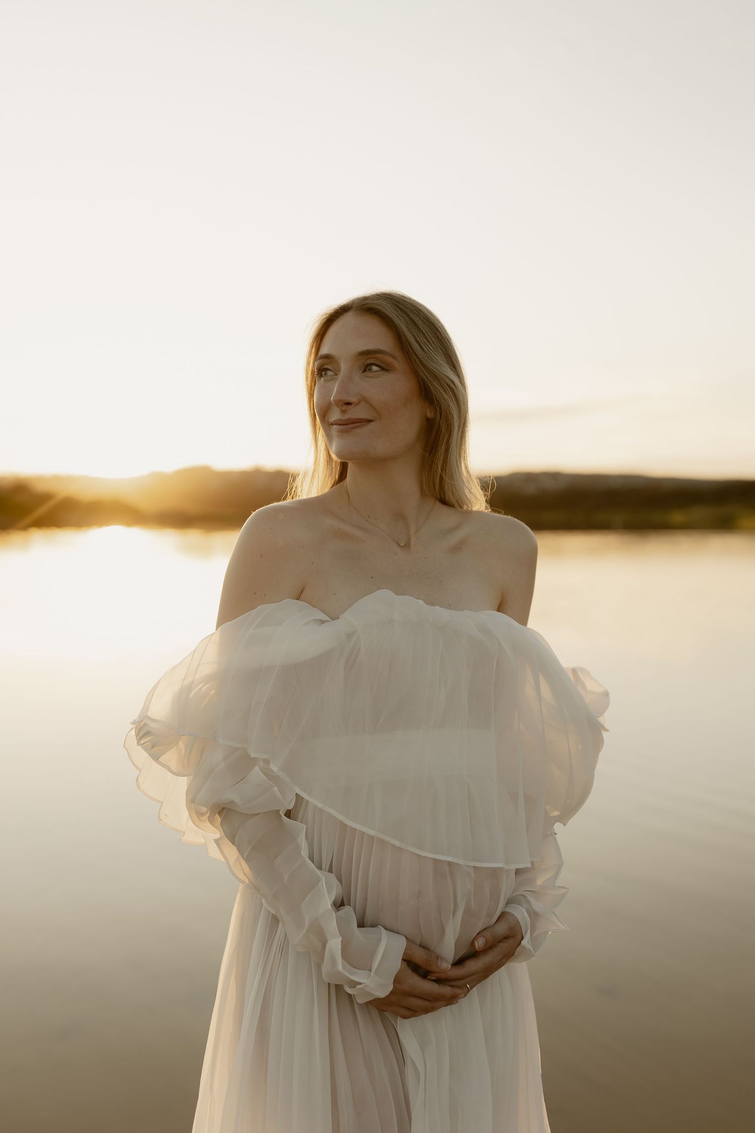 A woman in a flowing white dress poses gracefully by a tranquil lake at sunset.
