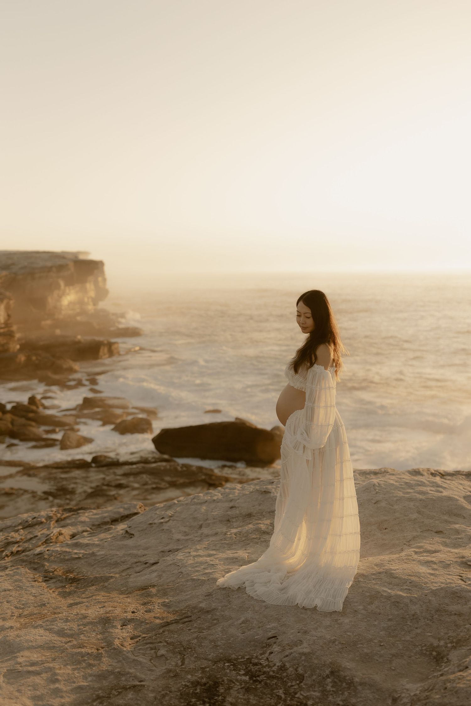 A figure in a flowing white dress stands on rocky coastal cliffs at golden hour overlooking the misty ocean waves.