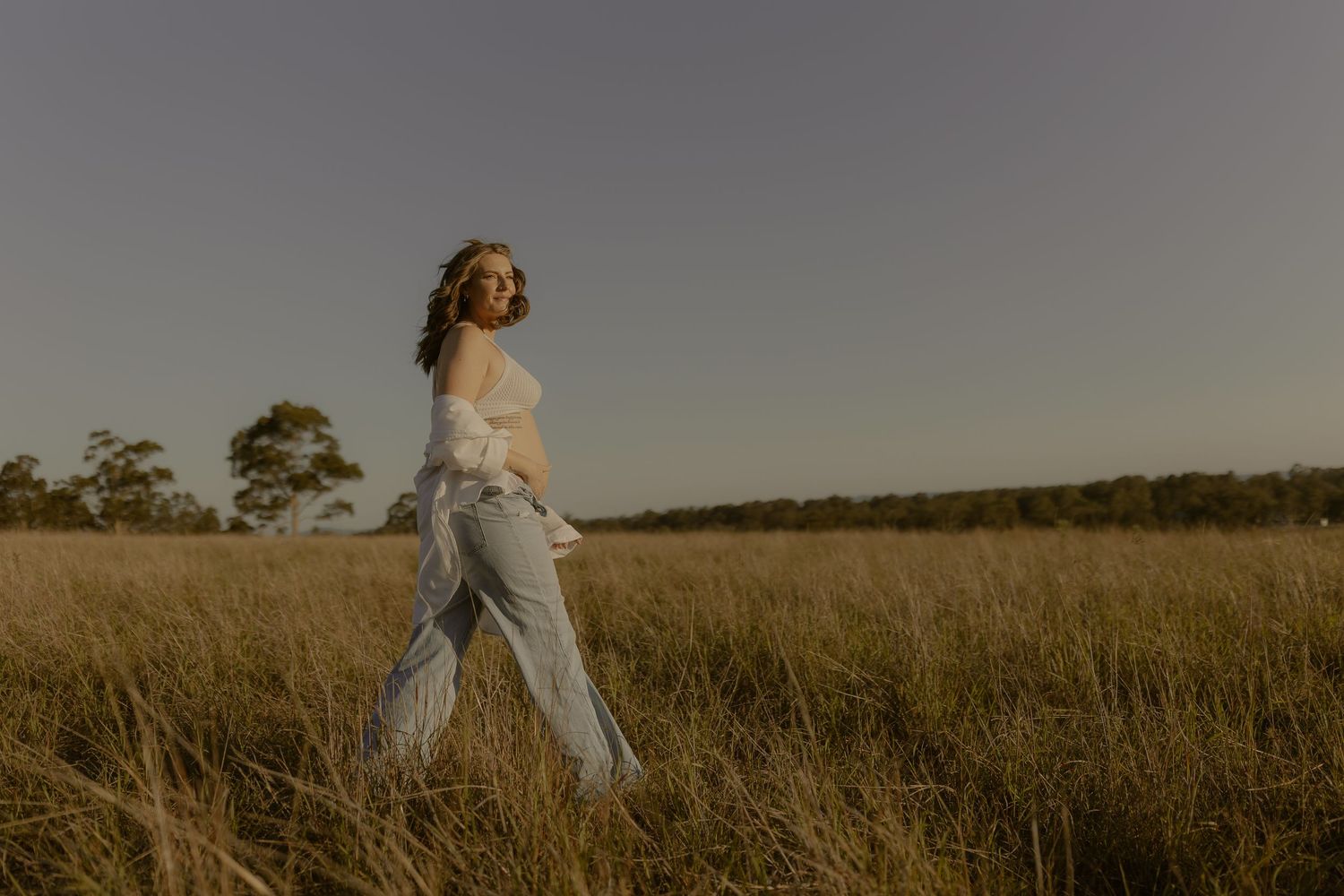 Person in light colored top walking through tall golden grass field at sunset.