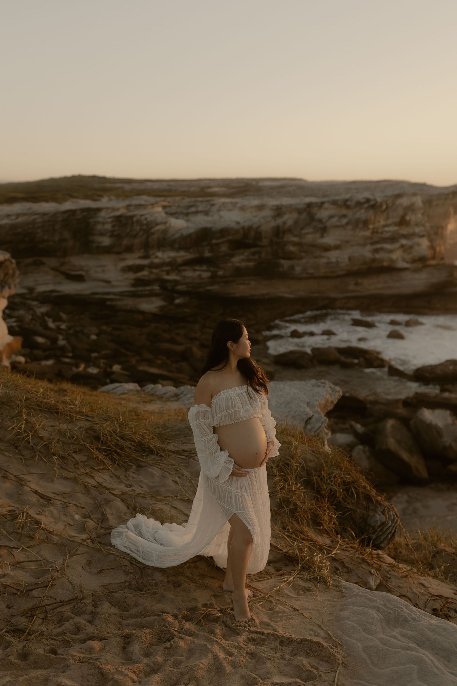 A woman in a flowing white dress stands on a coastal cliff during golden hour, overlooking a dramatic rocky shoreline.