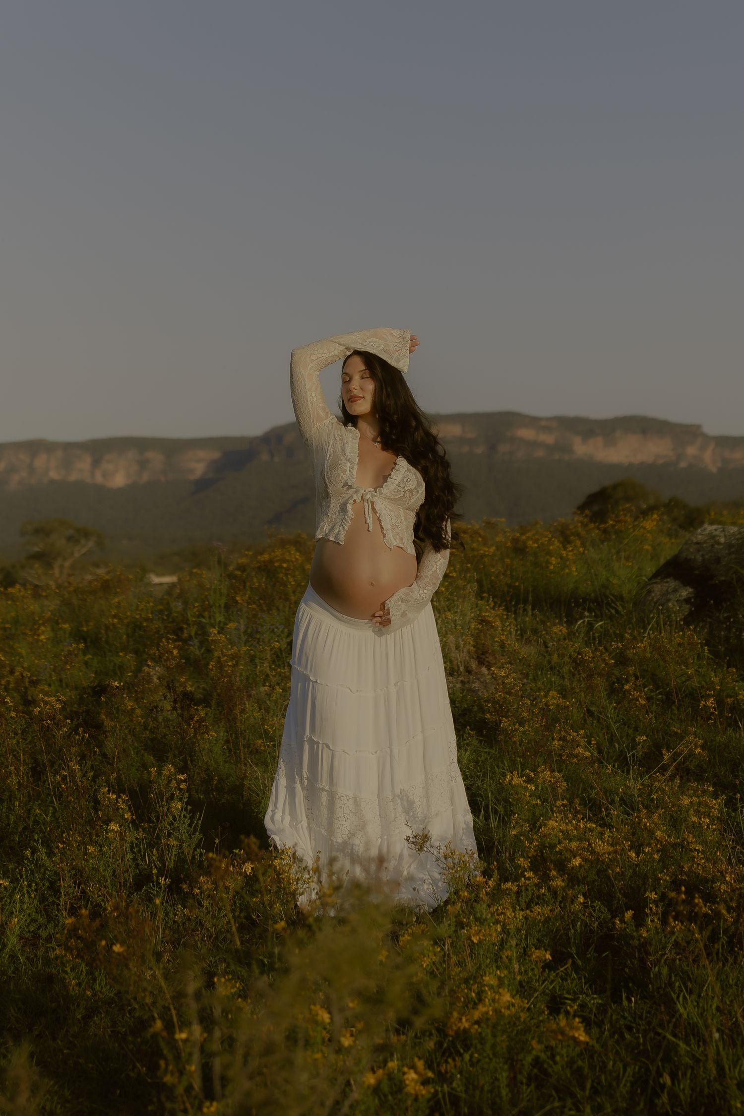 A person in a flowing white dress stands in a wildflower meadow with mountains visible in the background at sunset.