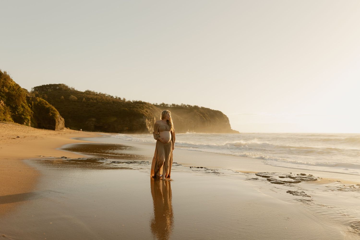 Pregnant woman and partner standing together at the shoreline during a warm golden sunset beach shoot.
